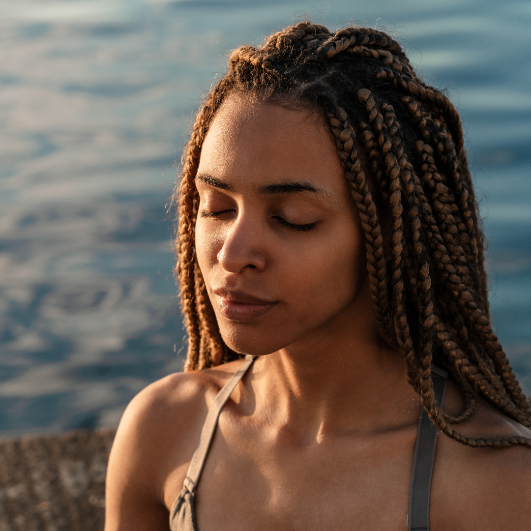 A young woman with brown braided hair standing near water with her eyes closed.
