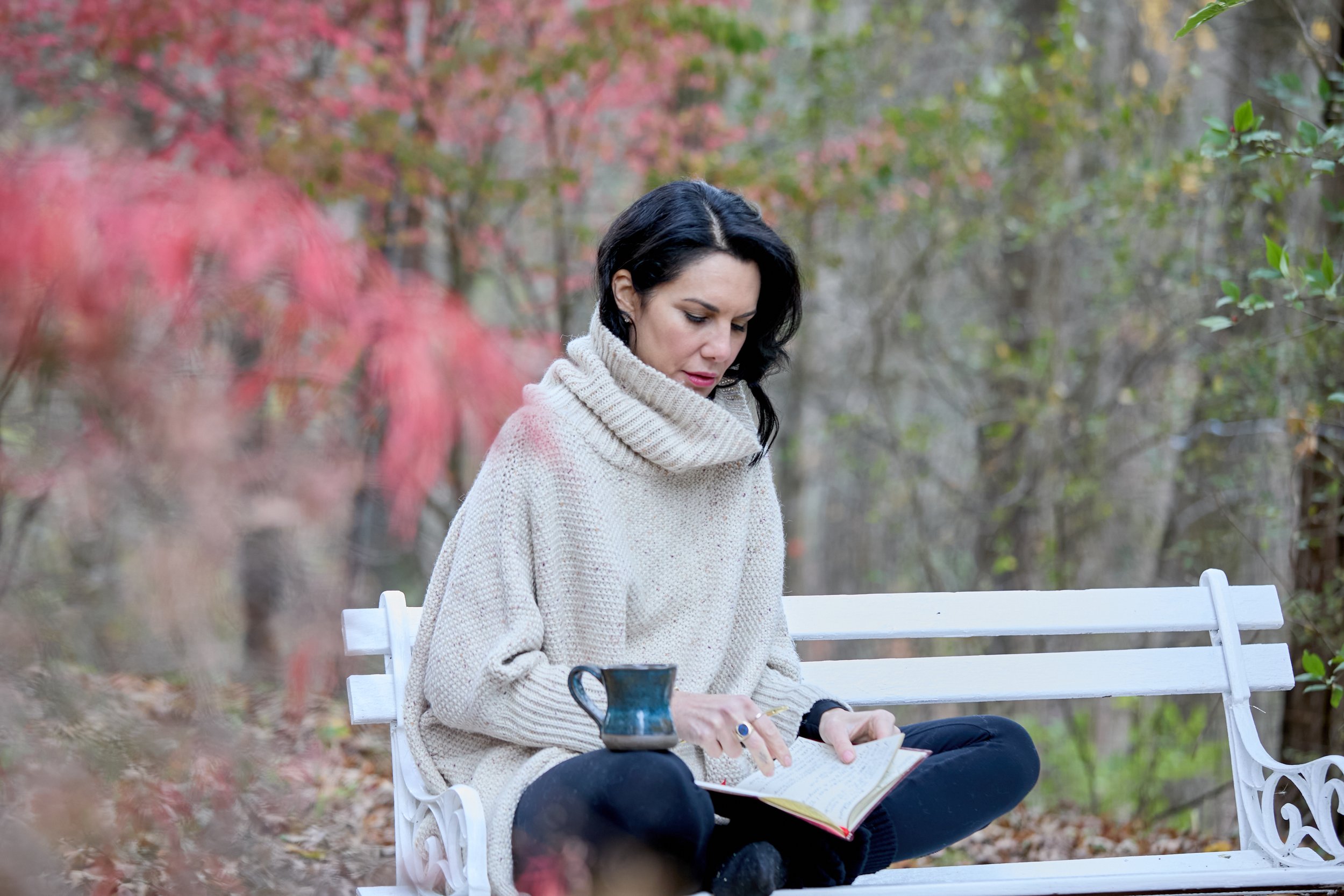 A woman with black hair, wearing a beige chunky knit sweater, is sitting cross-legged on a white park bench reading a book in a wooded area during autumn.