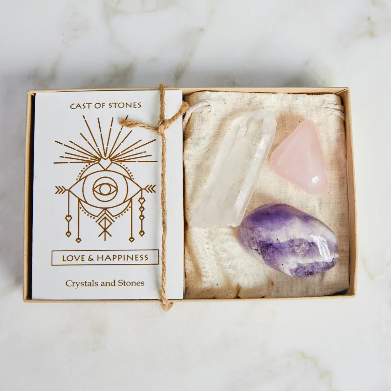 A gift box containing three polished crystals: clear quartz, rose quartz, and amethyst, with a card labeled 'Love & Happiness Crystals and Stones' and decorative elements on a white marble surface.