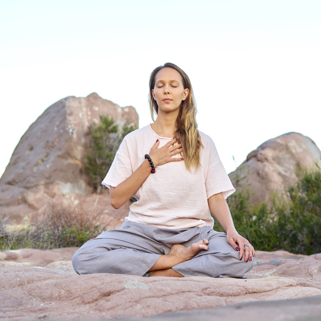 Woman practicing meditation outdoors, sitting cross-legged with one hand on her chest and the other on her knee, in a desert landscape with rocks and shrubbery.