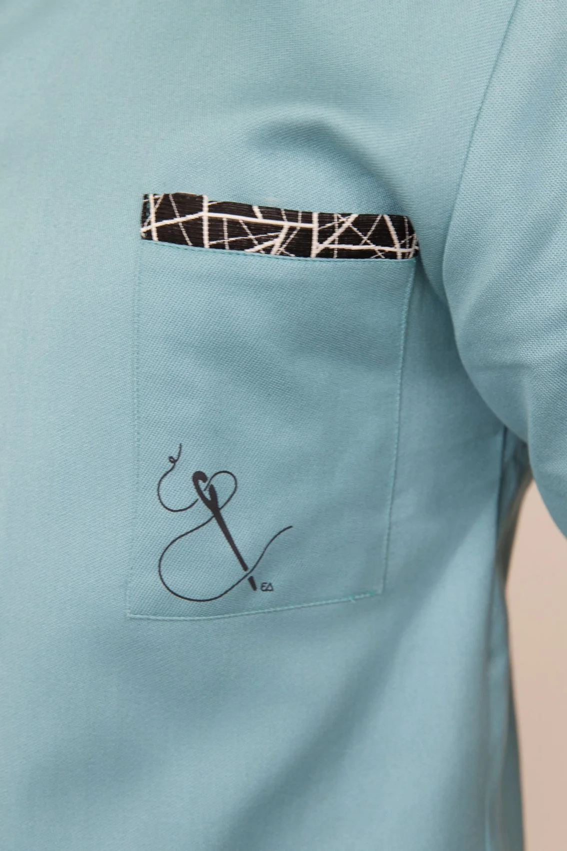 Une blouse médical turquoise avec une poche sur la poitrine, ornée d'un motif noir et blanc et d'un logo en forme de fil dentaire avec une aiguille.