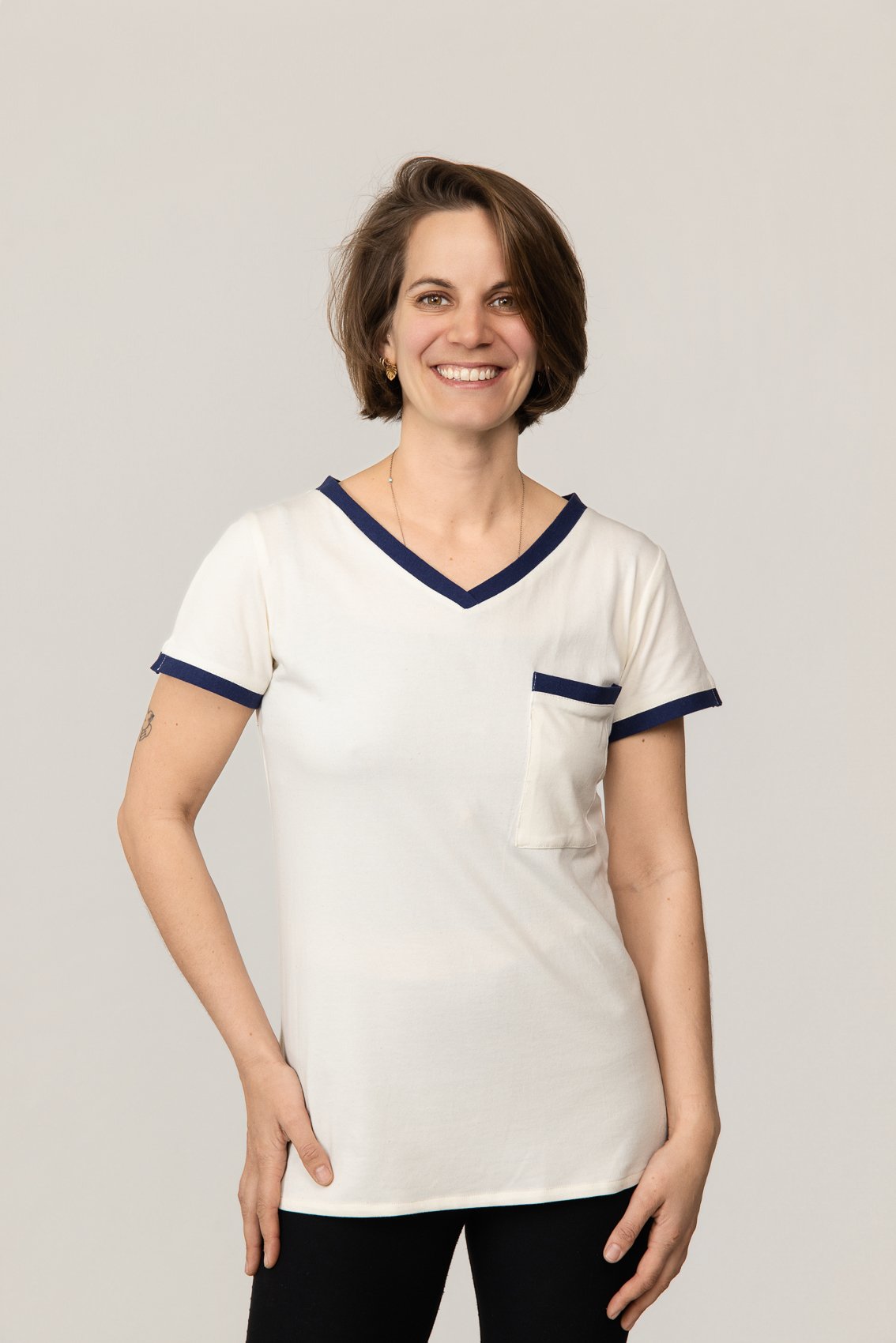 Femme souriante portant un t-shirt médical blanc à bordure bleu et une poche à la poitrine, posant contre un fond neutre.