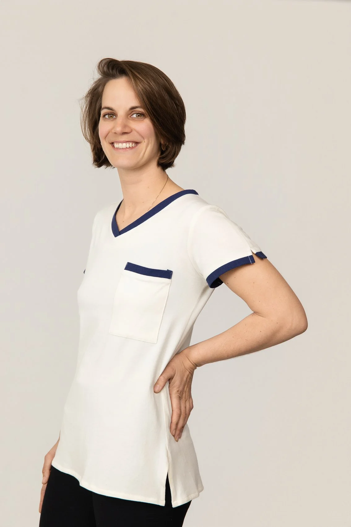 Femme souriant, portant un t-shirt médical blanc avec des accents bleus, posé de profil avec la main sur la hanche, fond blanc.