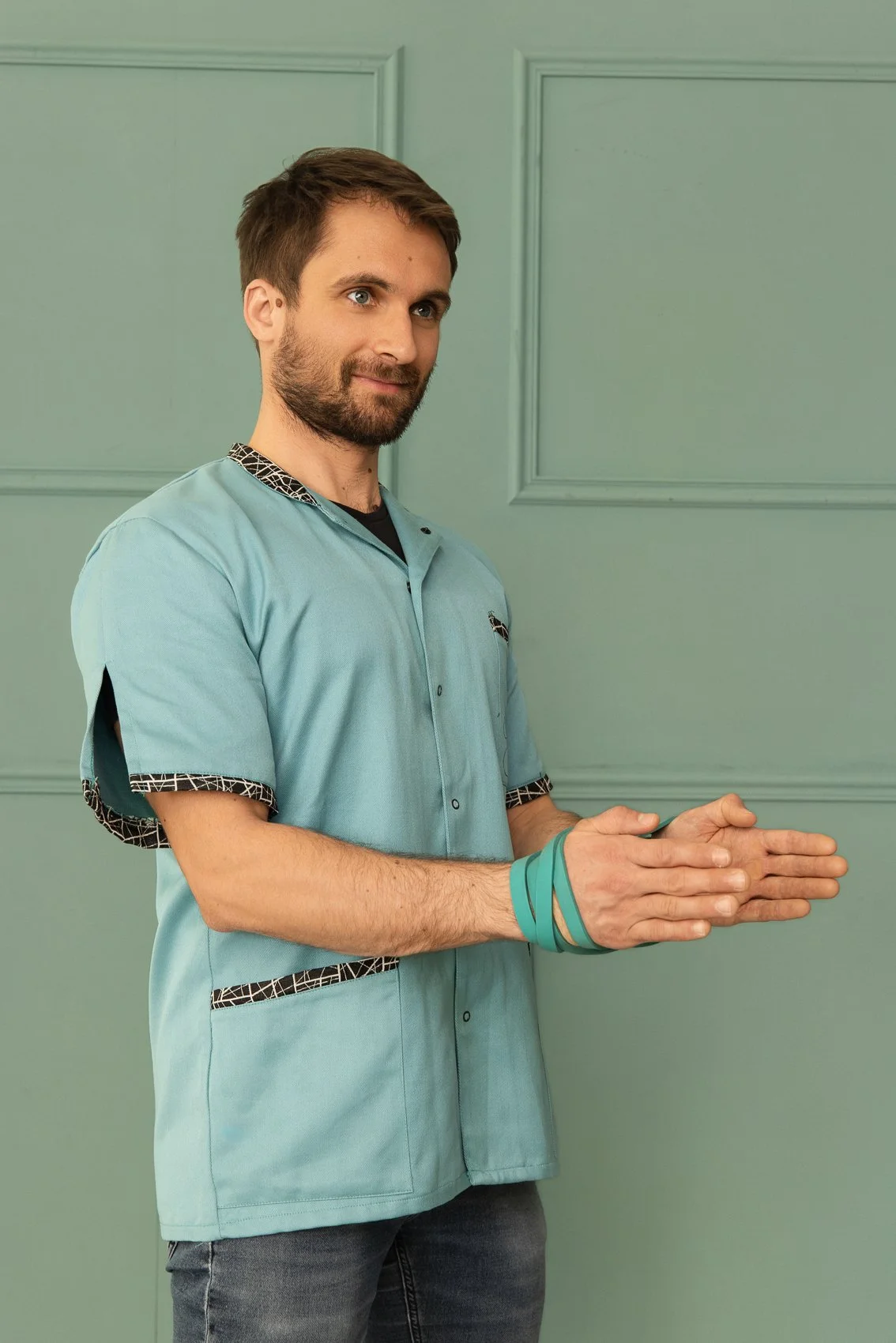 Homme en blouse médicale turquoise, mains jointes, portant des bracelets, devant un mur vert