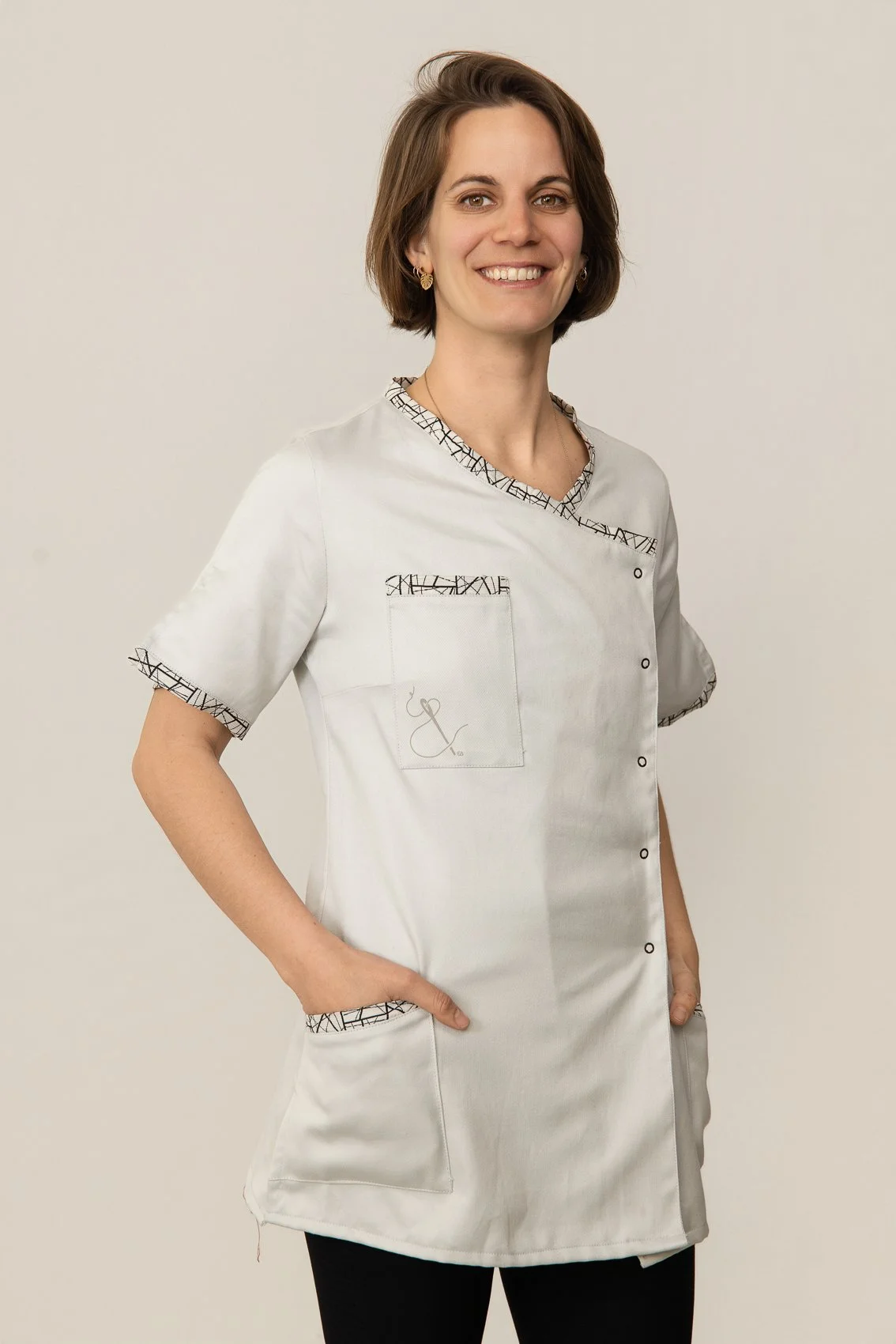Une femme portant une blouse médicale blanche avec des détails noirs, souriant, avec des cheveux bruns courts, debout contre un fond neutre.