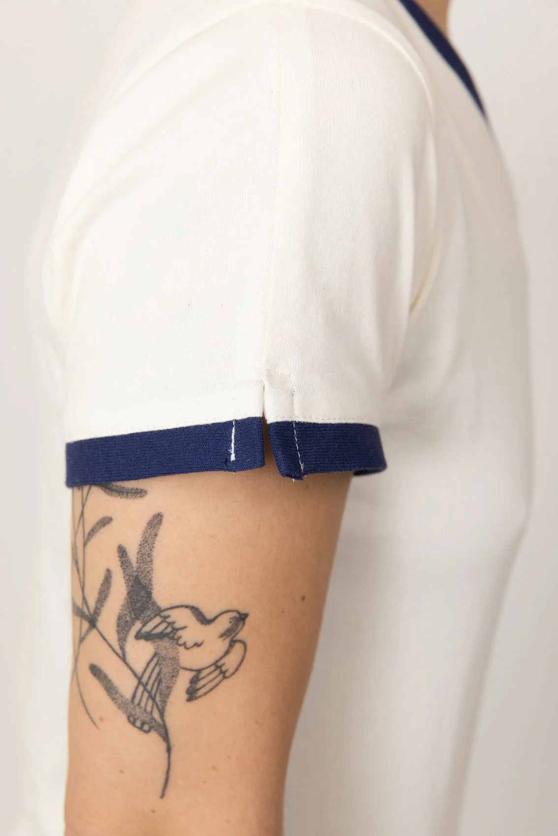 Bras gauche visible, vêtu d'un t-shirt médical blanc avec des bords bleus. Tatouage d'un oiseau en noir sur le bras nu.