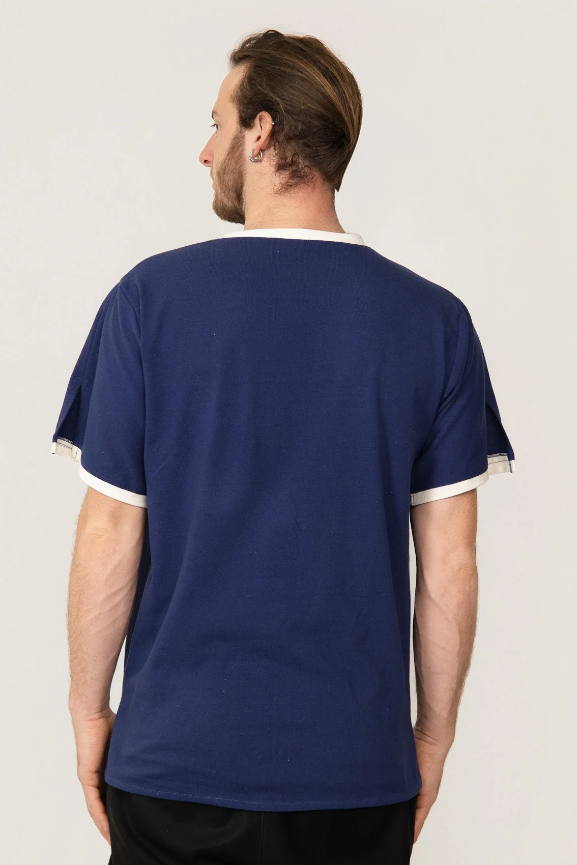 Vue de dos d'un homme portant un t-shirt médical bleu avec des bords blancs, avec ses cheveux longs, bruns, et un anneau à l'oreille, contre un fond clair.