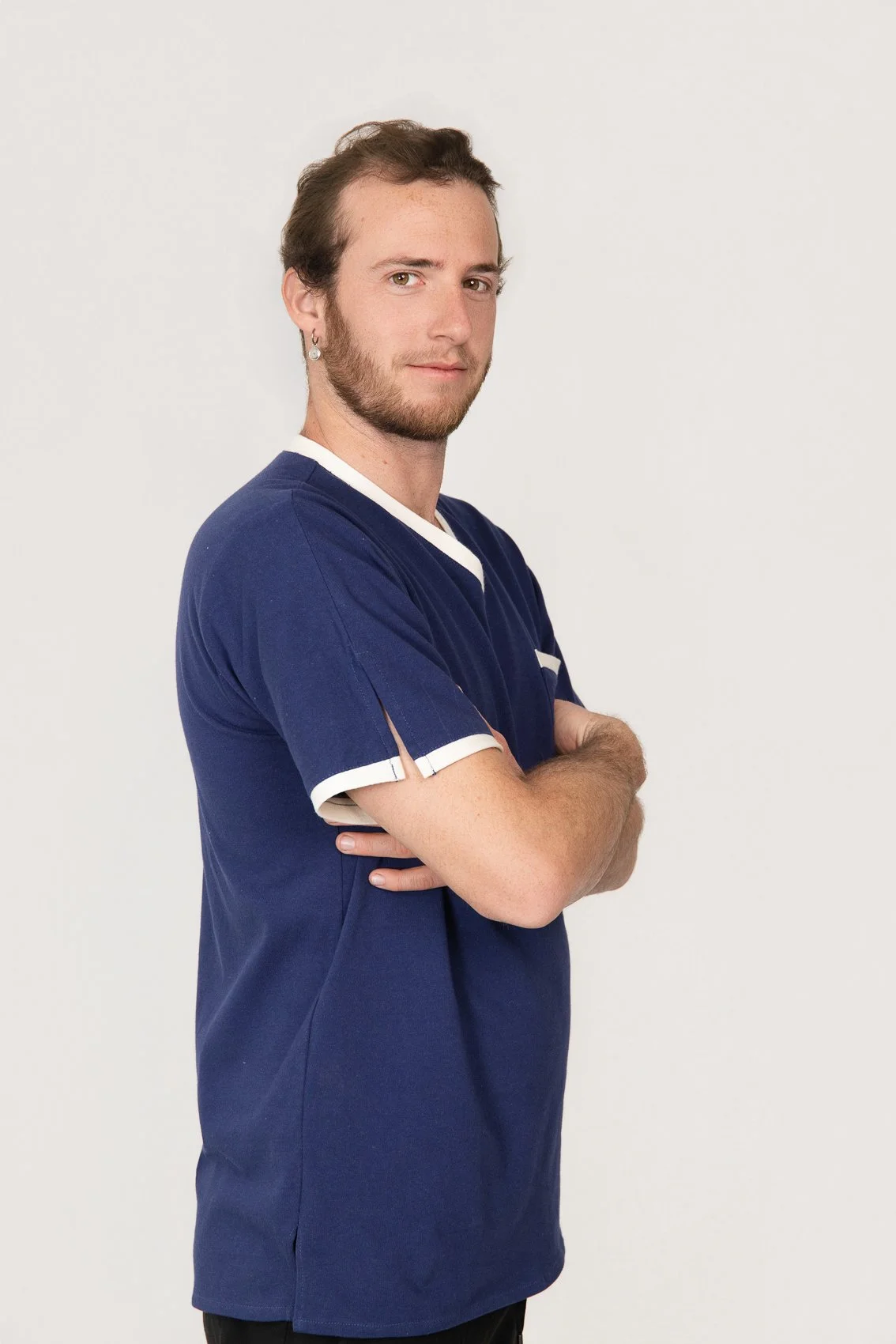 Homme avec barbe, portant un t-shirt médical bleu, croisant les bras, regardant vers la caméra, sur un fond blanc.