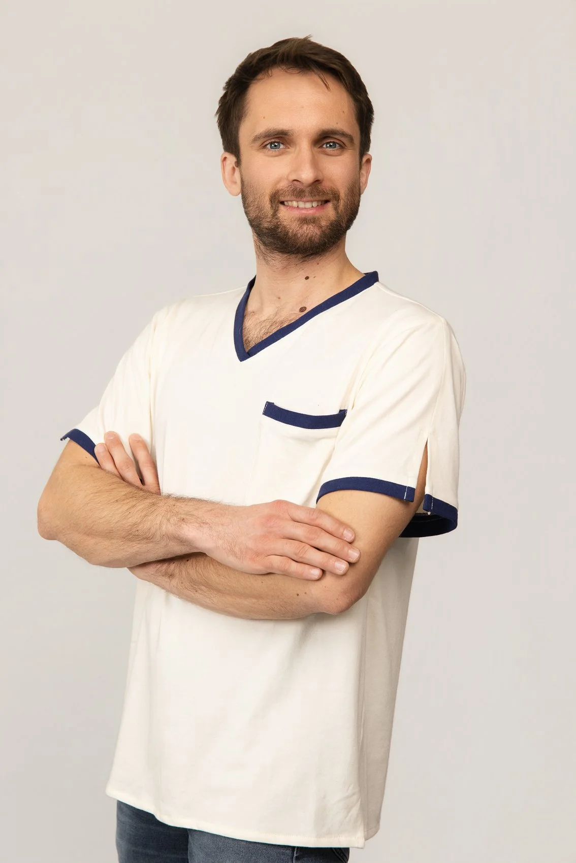Homme avec une barbe et un sourire, portant un t-shirt médical blanc avec des détails bleus, croisant les bras, debout contre un fond neutre.