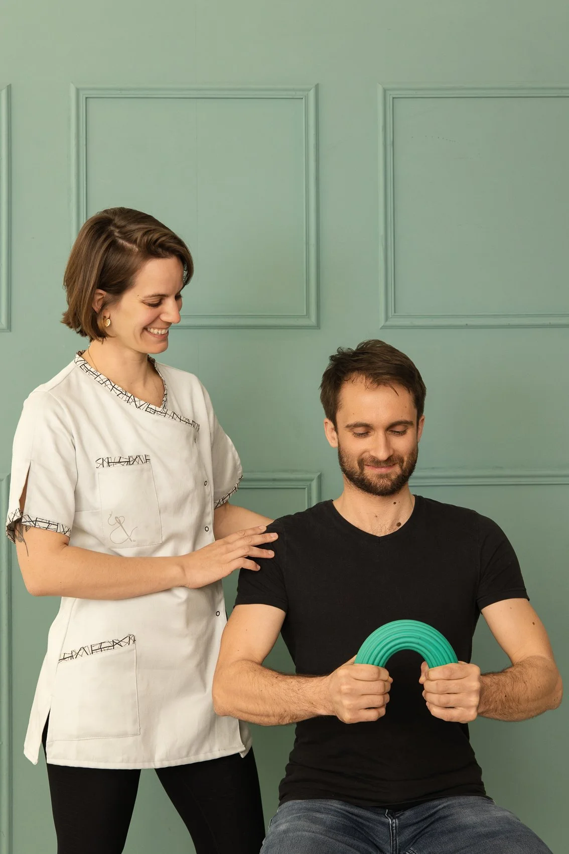 Une femme souriante, vêtue d'une blouse médicale blanche, touche l'épaule d'un patient assis tenant un flexbar, dans une pièce aux murs verts avec des moulures.