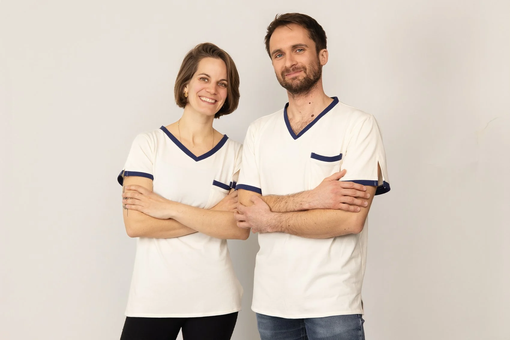 Deux personnes portant des t-shirt médicaux blancs avec des détails bleus, souriantes et croisées les bras, se tenant contre un mur blanc.