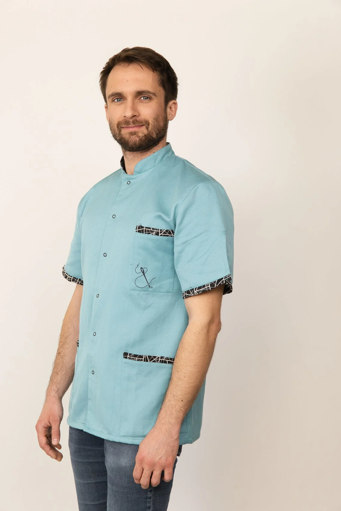 Homme en blouse médicale bleue avec détails noirs et blancs, debout contre un fond blanc, souriant légèrement.
