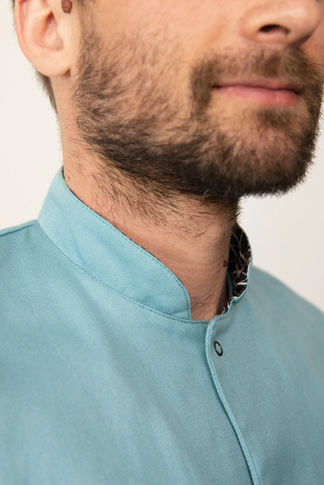 Zoom sur le cou et le menton d'un homme portant une blouse médicale bleu pâle, avec une barbe courte et soignée.