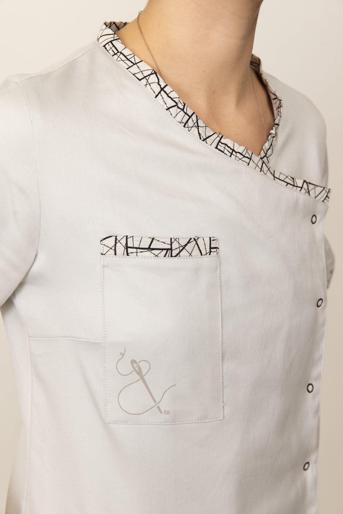 Une personne porte une blouse médicale blanche avec des détails de couture noirs et blancs à la collerette et à la poche. La personne porte également un collier fin.