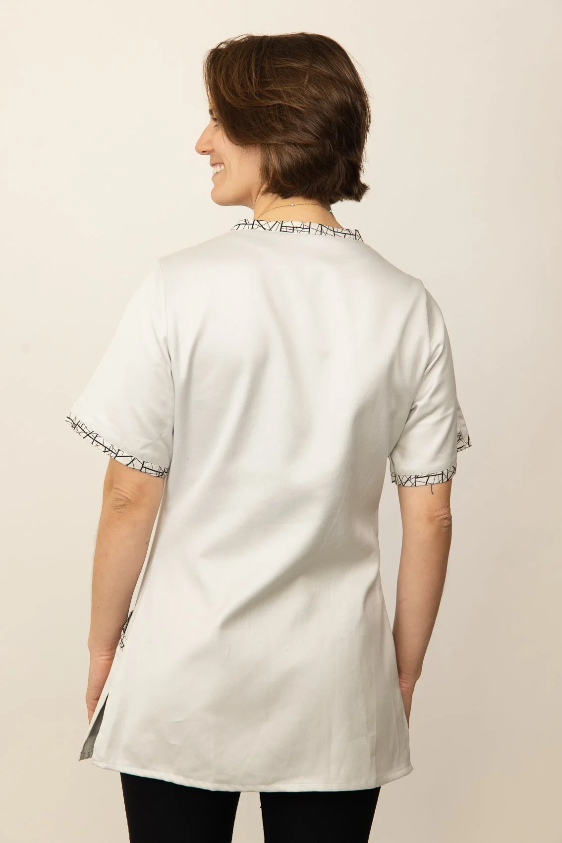 Femme souriante portant une blouse médicale blanche à manches courtes avec des détails à l'encolure et aux manches.