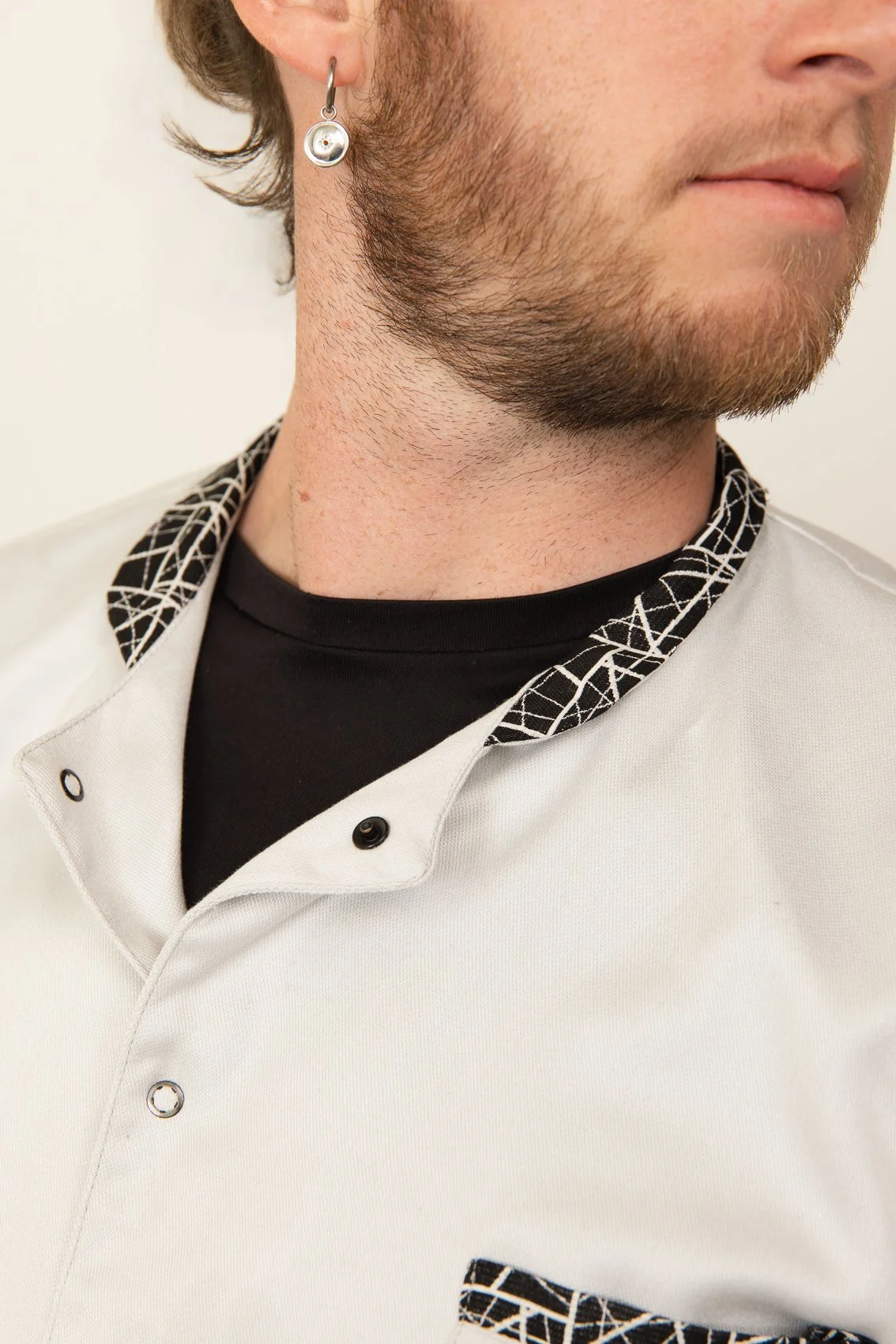 Un homme portant une blouse médicale blanche avec un col à motif géométrique noir et blanc, et un t-shirt noir en dessous. Il porte une boucle d'oreille en forme de cercle.