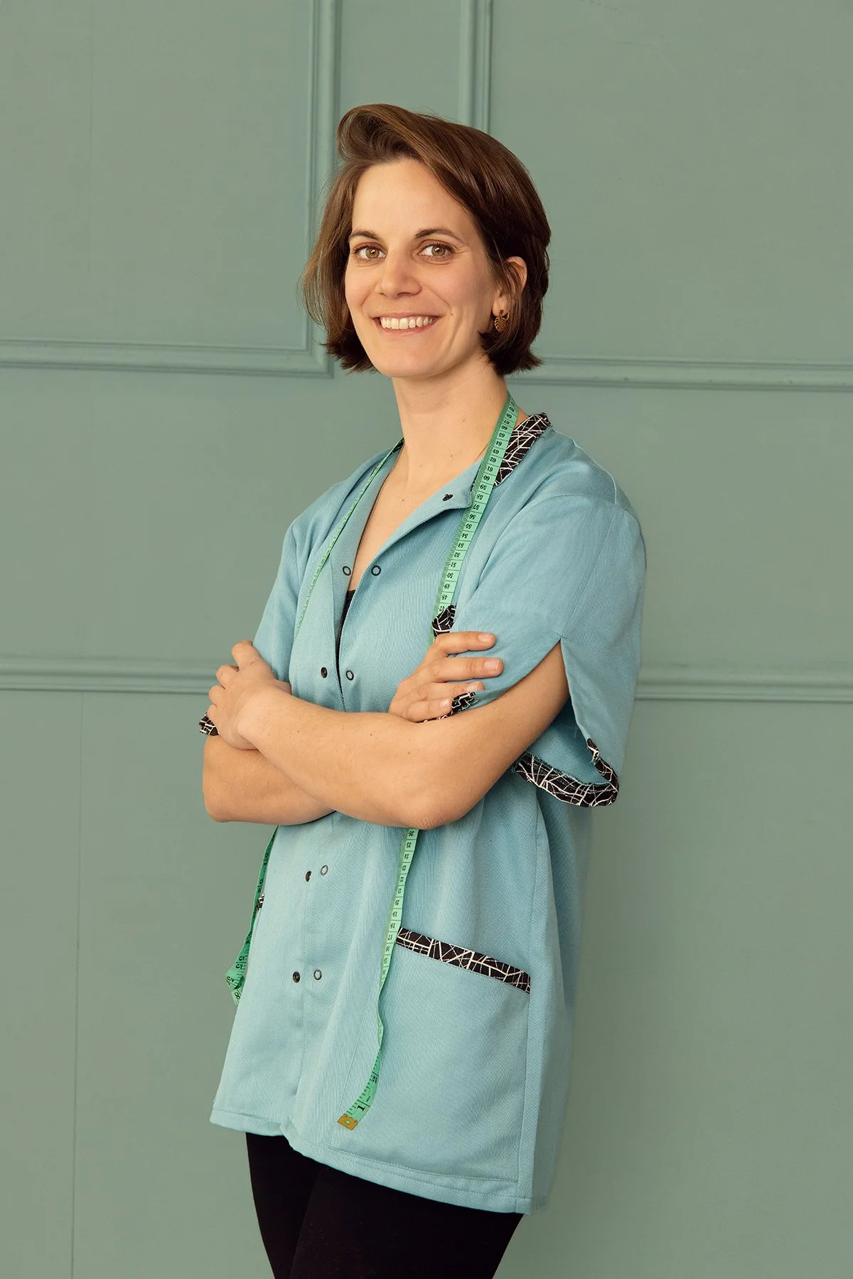 Une femme habillée en blouse médicale bleue avec un ruban de mesure autour du cou, souriant, avec un fond vert.