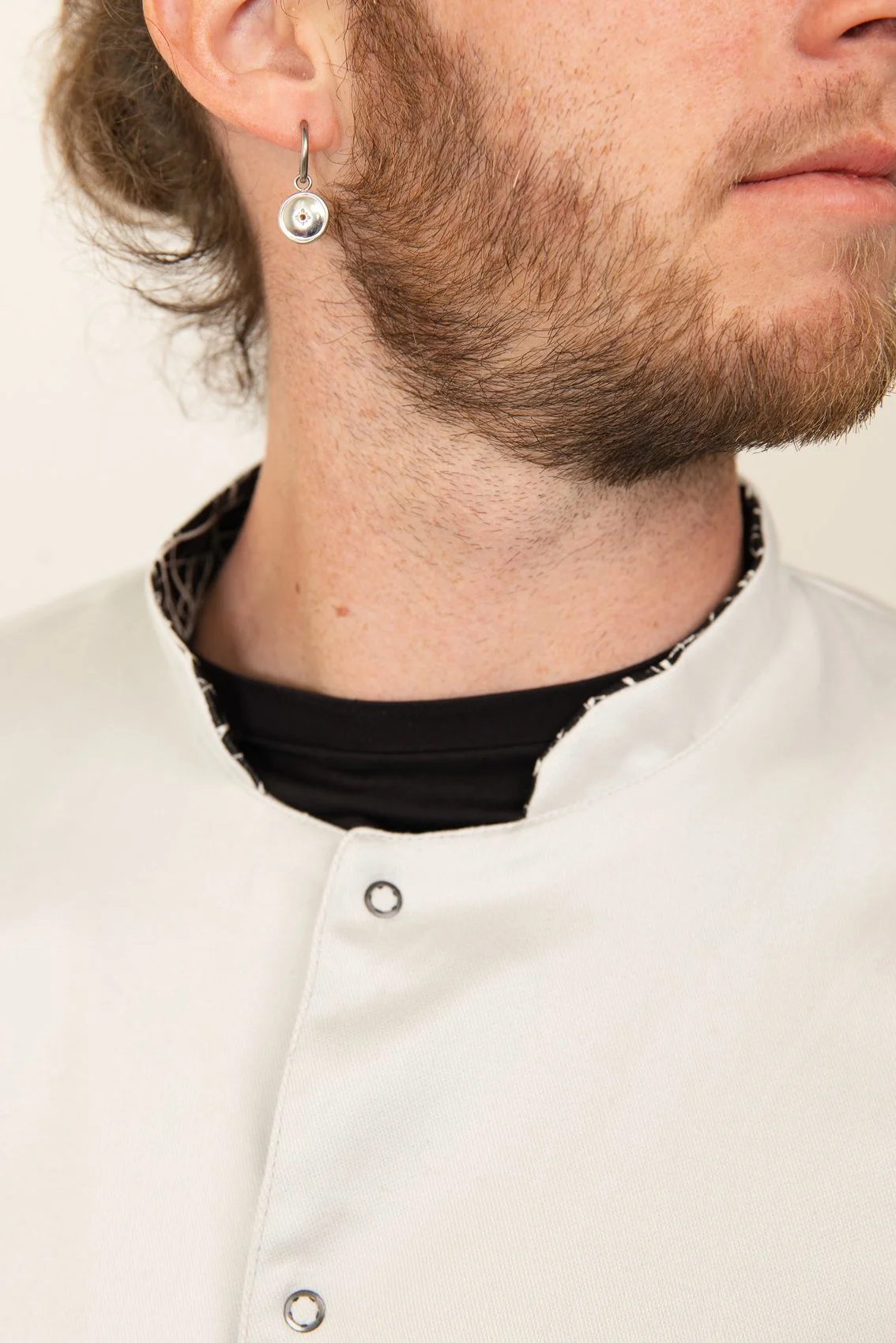 Close-up du visage d'un homme portant une blouse médicale blanche à col mao et un cercle d'oreille en argent, avec une barbe légère et des cheveux bouclés
