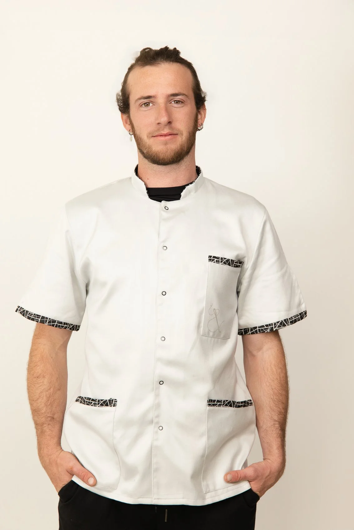 Jeune homme portant une blouse médicale blanche de chef, debout face à un mur blanc, main dans les poches.