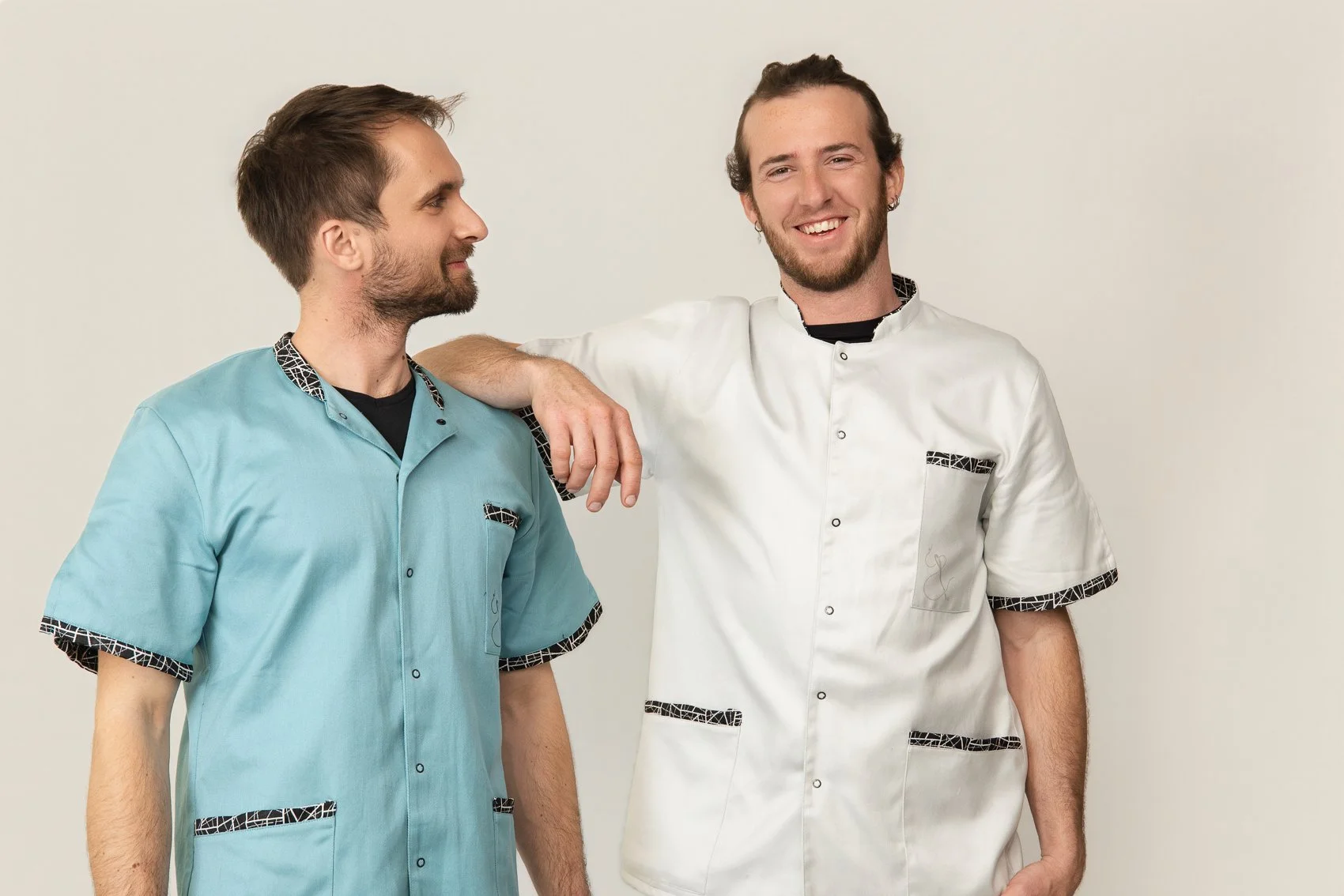 Deux hommes souriants portent des blouses médicales de couleur différente, l'un en bleu et l'autre en blanc, avec des détails en motif noir et blanc, se tenant contre un mur blanc. L'un pose sa main sur l'épaule de l'autre.