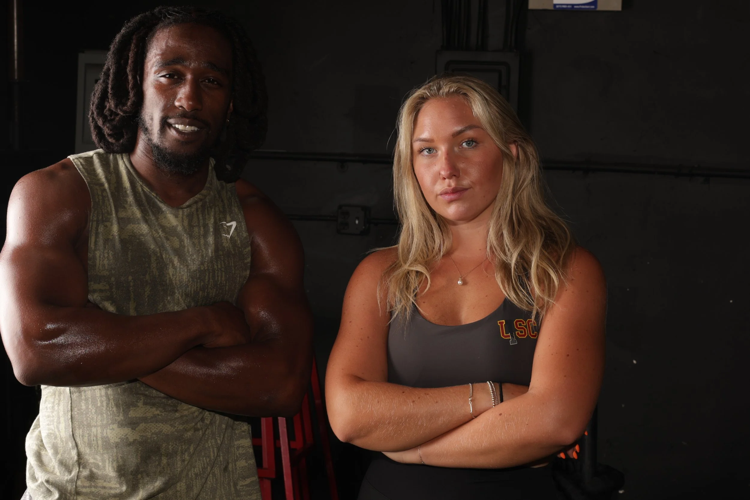 Two athletes, a man with dark skin and dreadlocks wearing a sleeveless shirt, and a woman with light skin and blonde hair wearing a sleeveless top with LSCO logo, standing side by side with their arms crossed in a dark indoor setting.