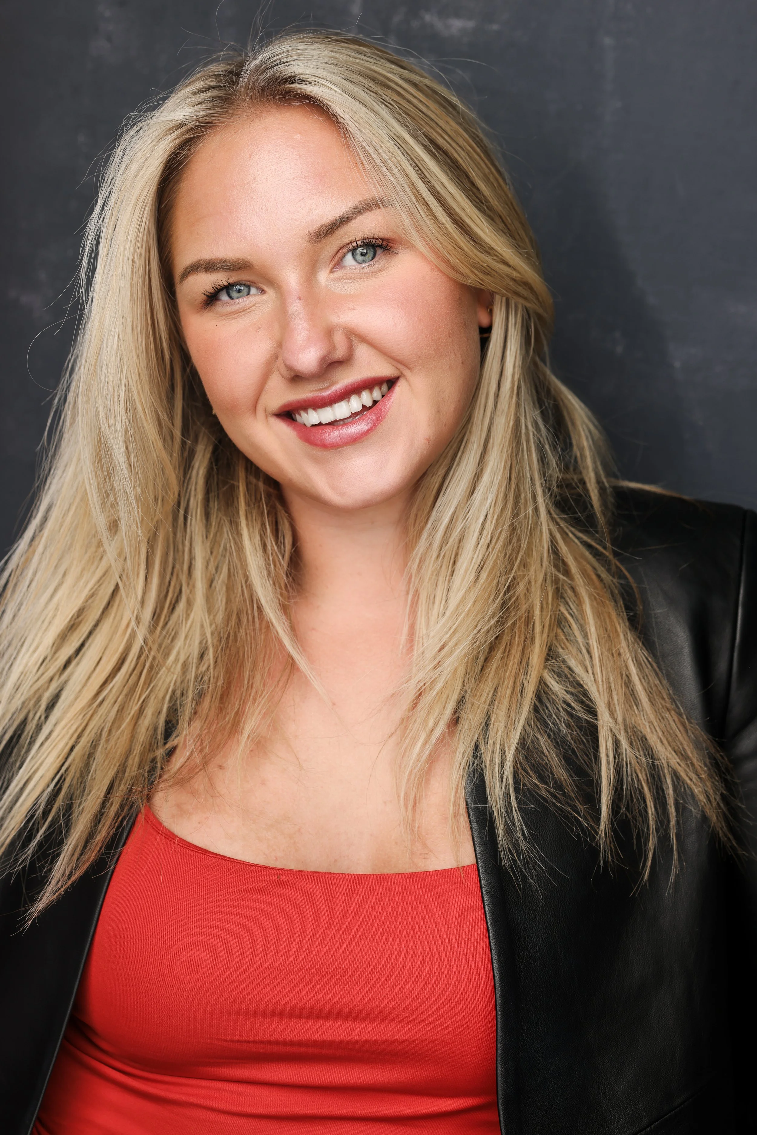 A woman with long blonde hair, blue eyes, and a bright smile, wearing a red top and a black jacket, stands against a dark background.