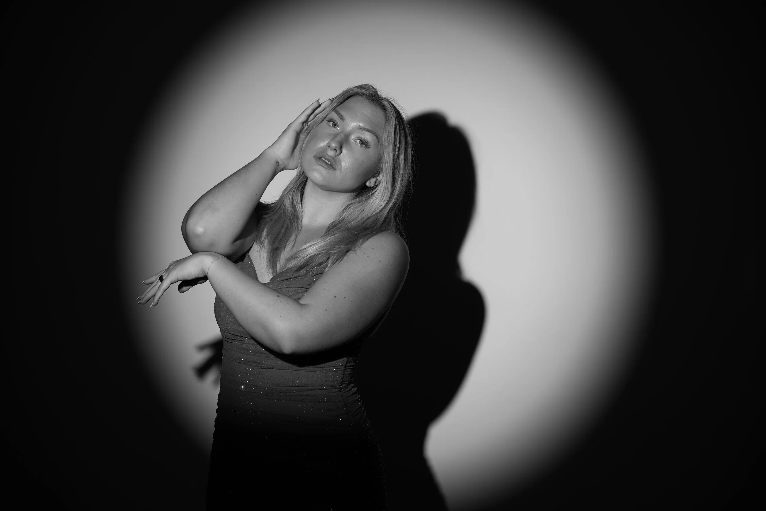 Black and white photo of a woman with long hair, posing with one hand on her head and the other arm crossed over her chest, with soft lighting and a shadow cast behind her.