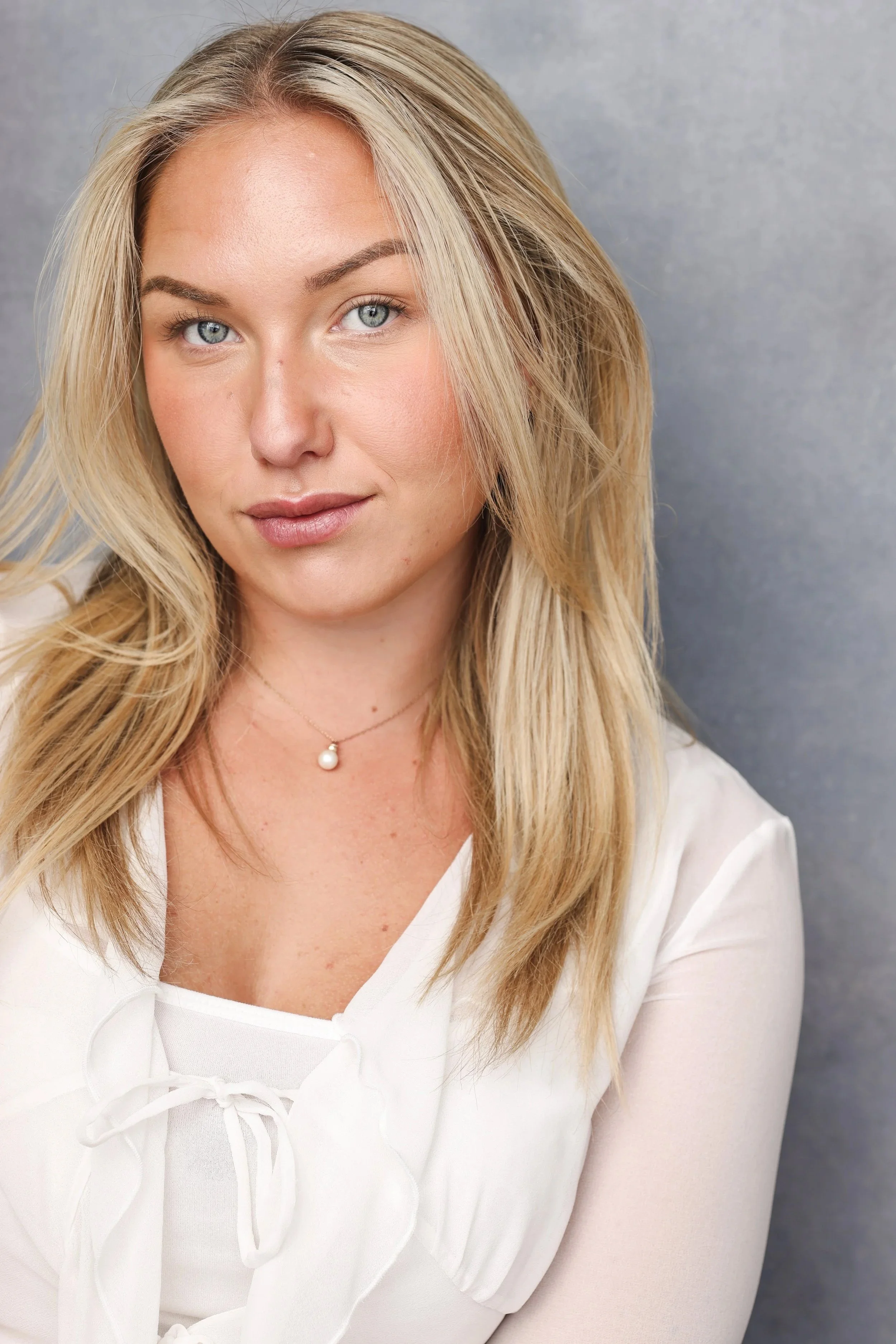 Close-up portrait of a young woman with blonde hair and blue eyes, wearing a white blouse with a bow and a pearl necklace, against a textured gray background.