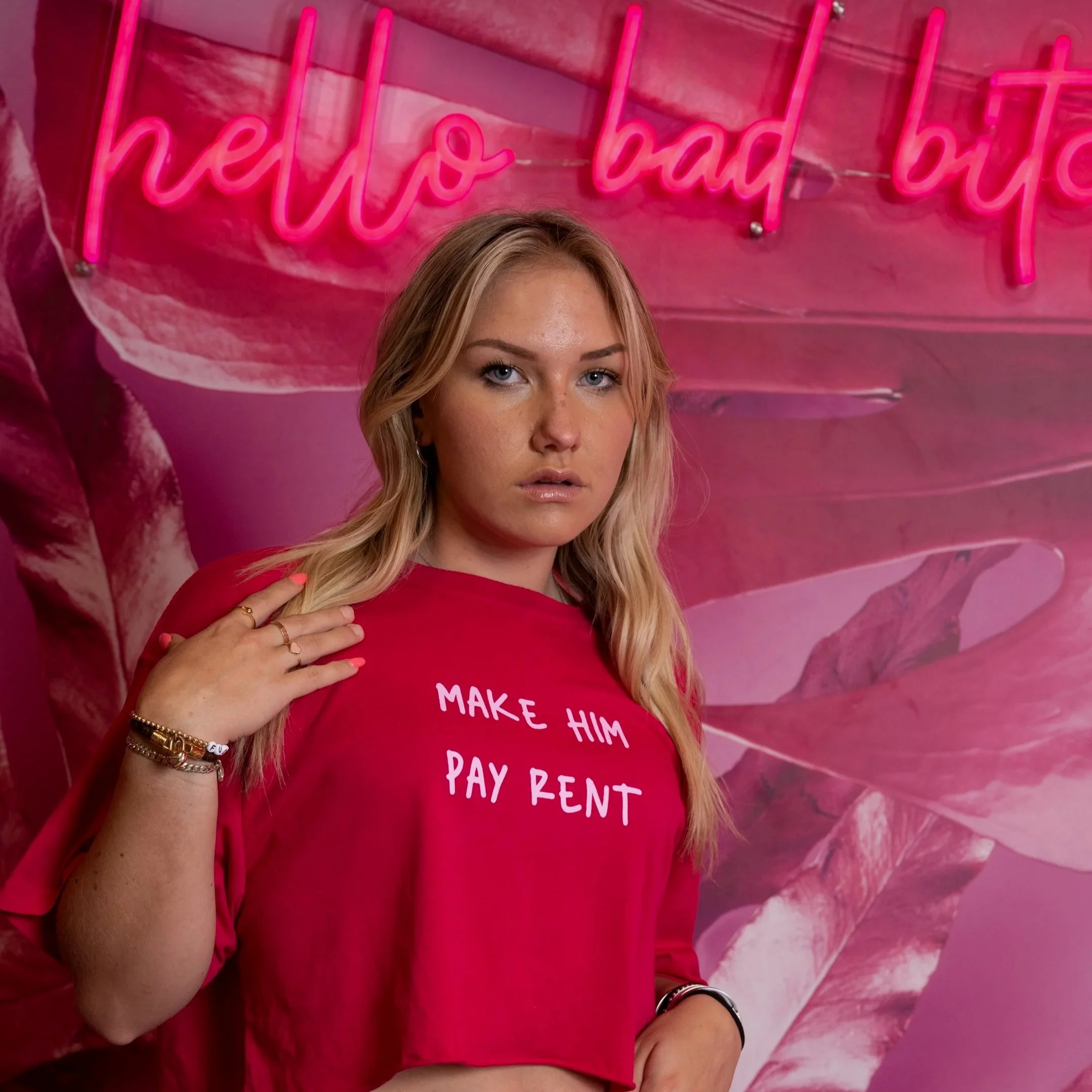 A young woman with blonde hair, wearing a pink T-shirt that says 'MAKE HIM PAY RENT,' is standing in front of a pink wall with a neon sign that says 'hello bad b***h' in cursive. She has a serious expression, and her left hand is touching her shoulde
