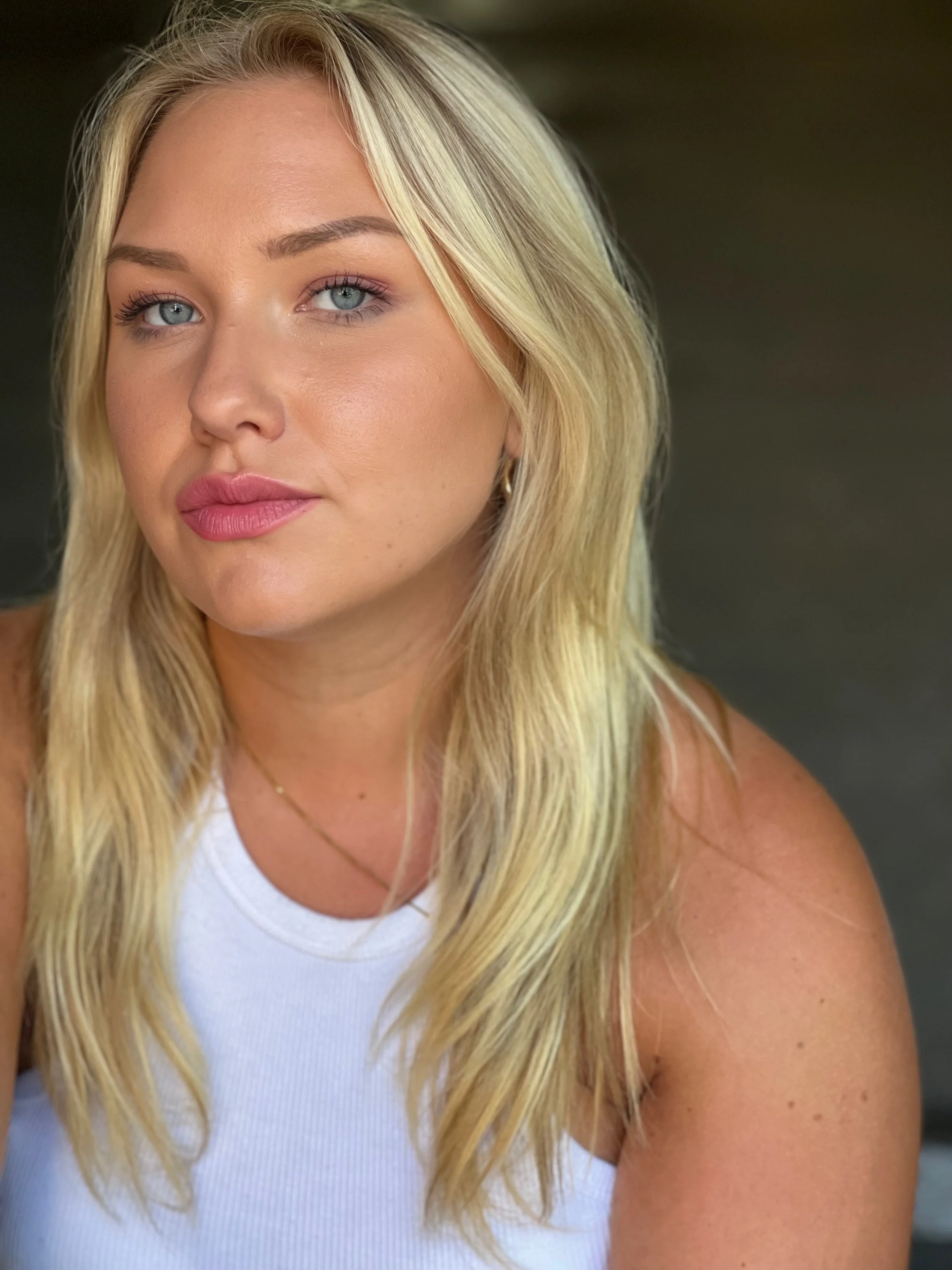A close-up of a woman with long blonde hair, blue eyes, wearing makeup, a white sleeveless top, and a thin gold necklace.