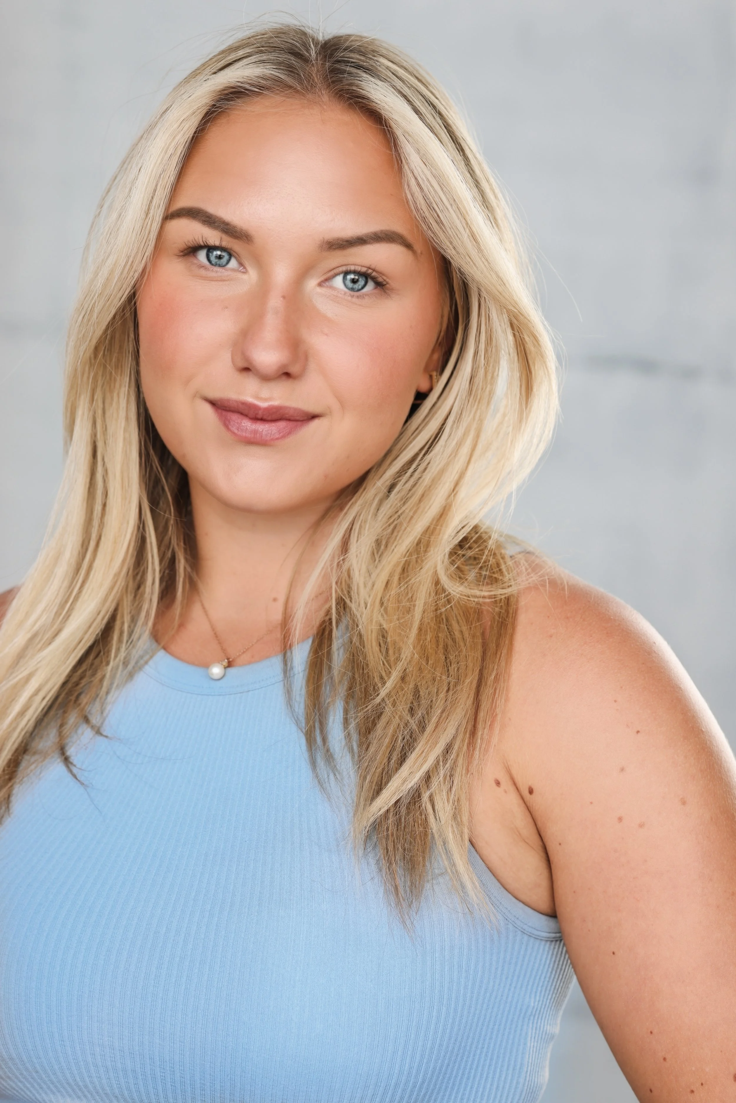 Portrait of a young woman with blonde hair, blue eyes, wearing a light blue top and a pearl necklace, smiling softly.