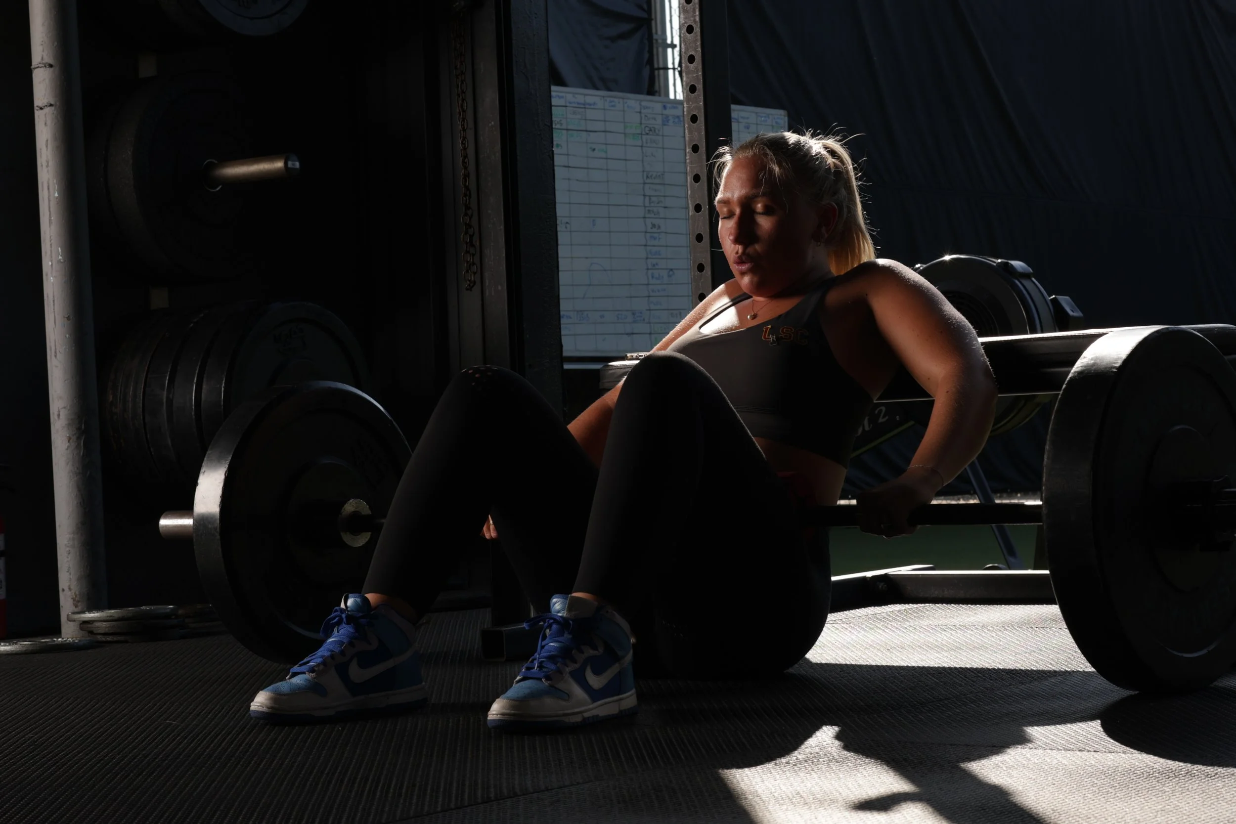 A woman wearing a sports bra and athletic shoes is doing a workout with a barbell on her hips in a gym, with sunlight contrasting the dark background.