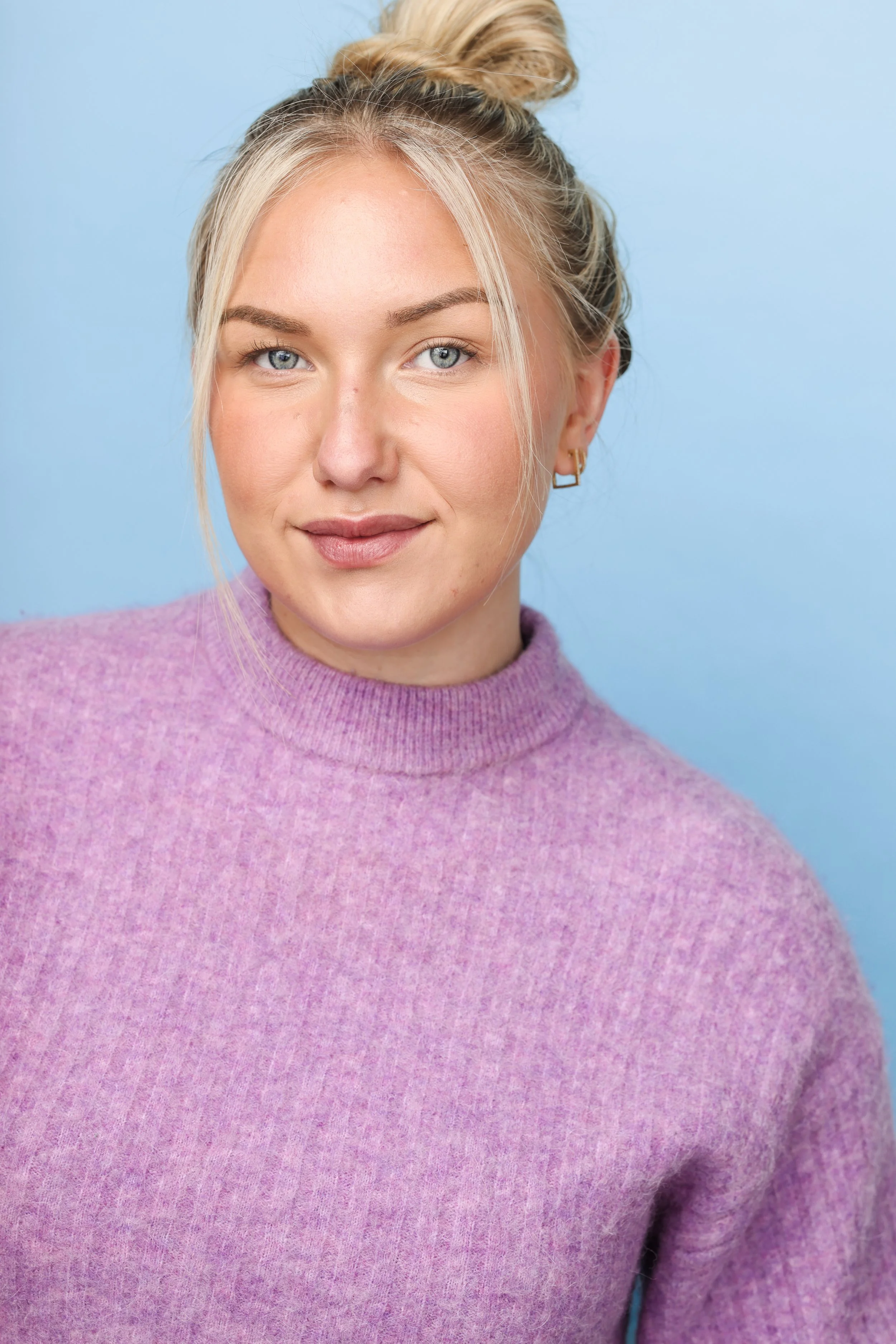 Close-up portrait of a young woman with blonde hair styled in a messy bun, blue eyes, wearing a purple turtleneck sweater, against a light blue background.