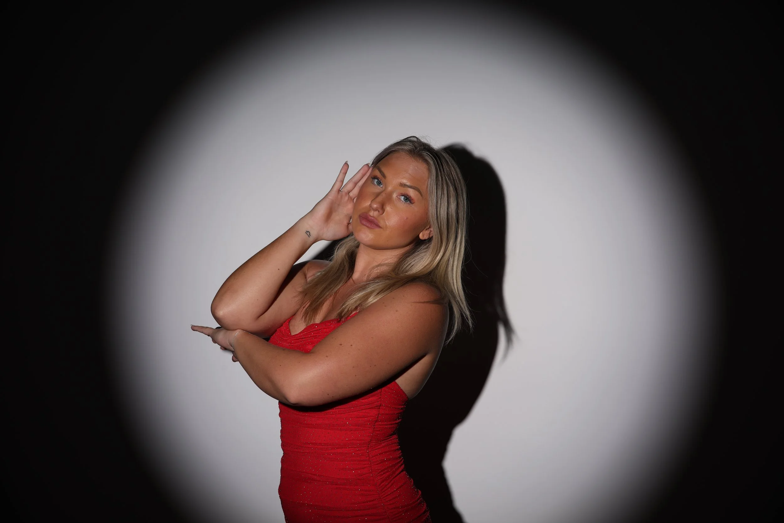 A woman with blonde hair in a red dress posing with her hand near her face against a white background with a circular black border.