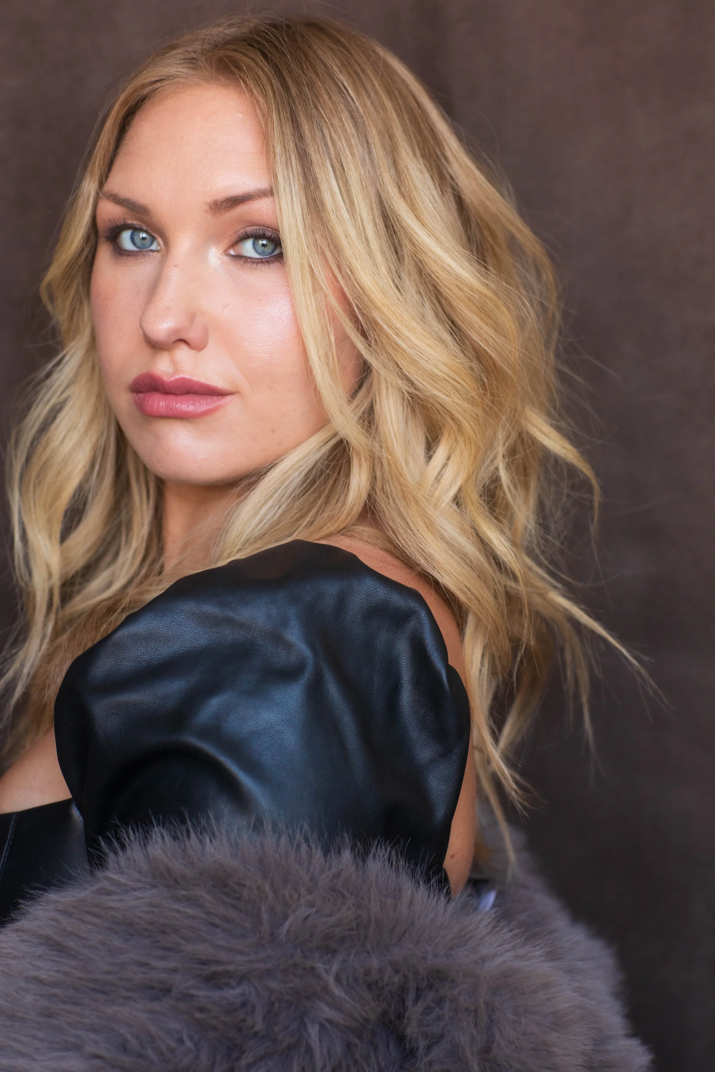 A woman with long blonde wavy hair and blue eyes wearing a black leather sleeve and a furry coat.