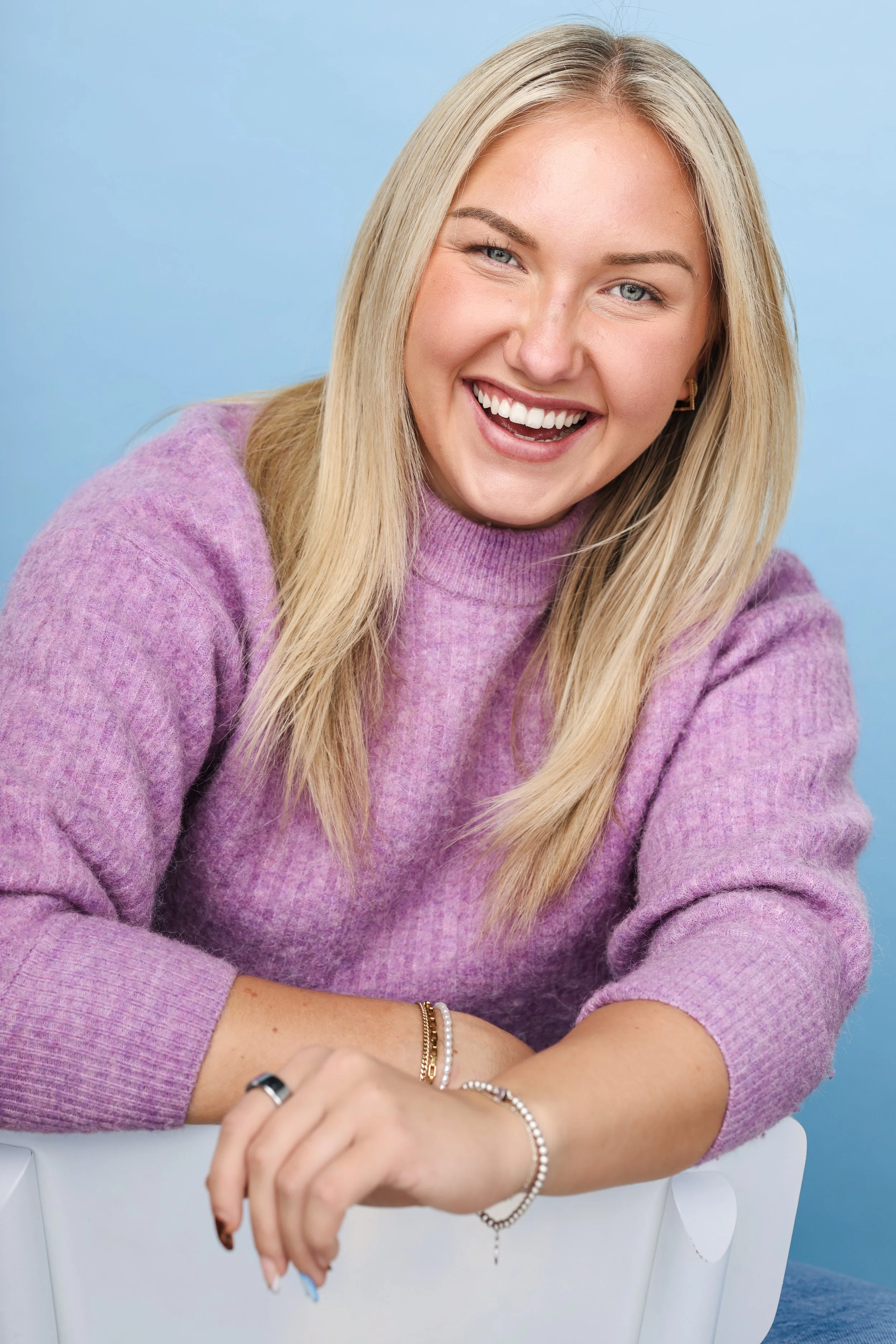 A smiling woman with blonde hair, wearing a purple sweater, sitting against a light blue background.