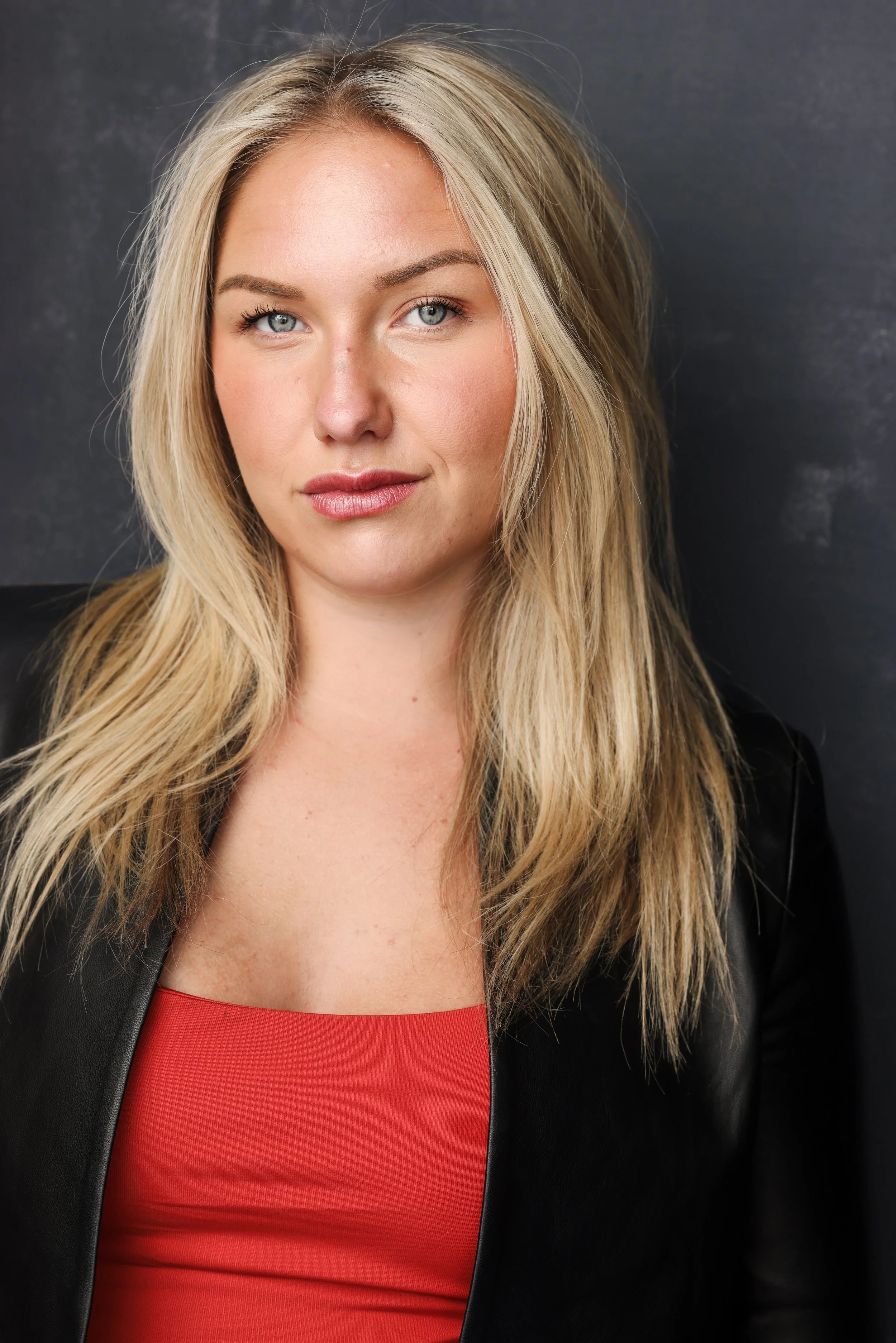 A woman with blonde hair, blue eyes, wearing a black jacket and red top, posing against a dark background.