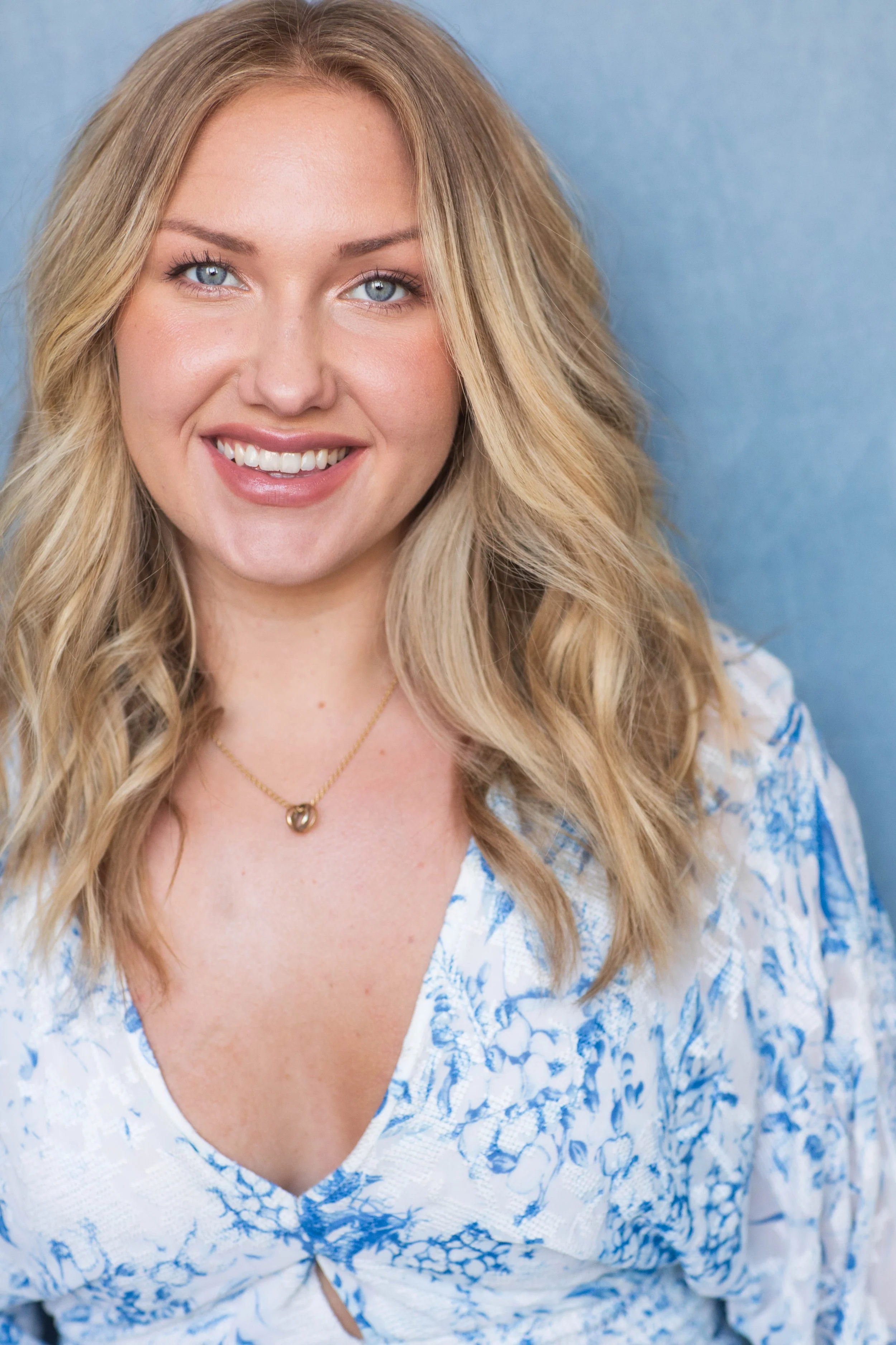 A smiling woman with long blonde hair, blue eyes, wearing a white and blue floral top, and a gold necklace, against a blue background.