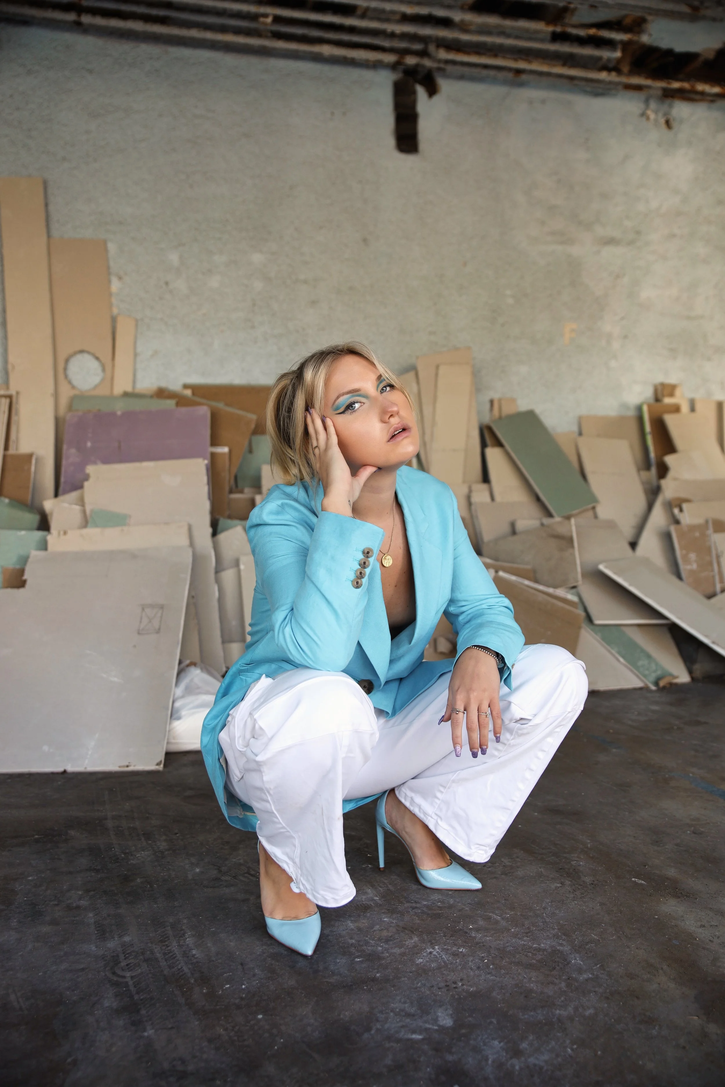 A woman in a blue blazer and white pants squats in front of a stack of drywall sheets in an unfinished or abandoned space. She is wearing blue high heels and has blonde hair with blue eye makeup, resting her head on her hand with a contemplative expr