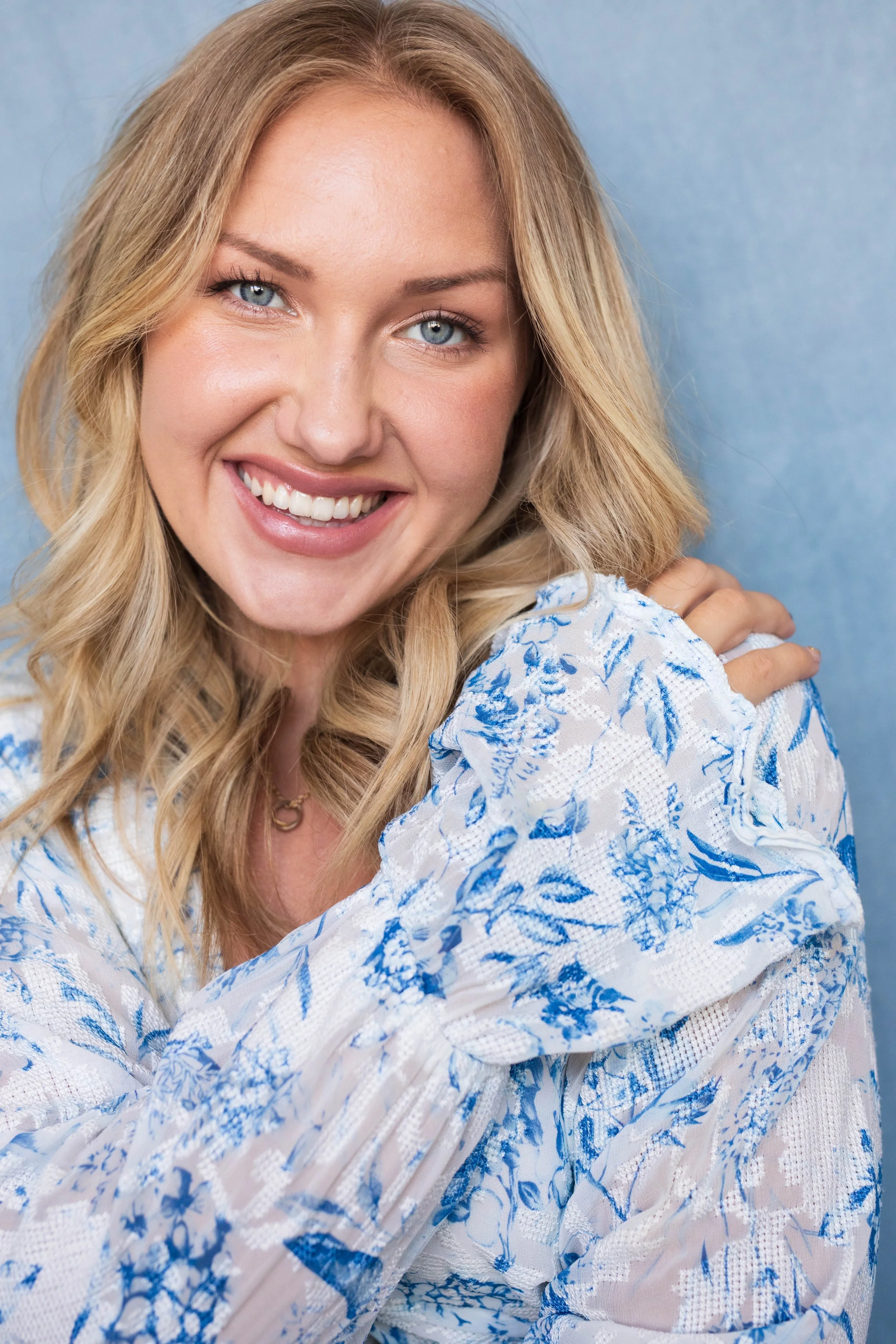 A smiling woman with blonde hair and blue eyes wearing a white blouse with blue floral patterns, posing against a light blue background.