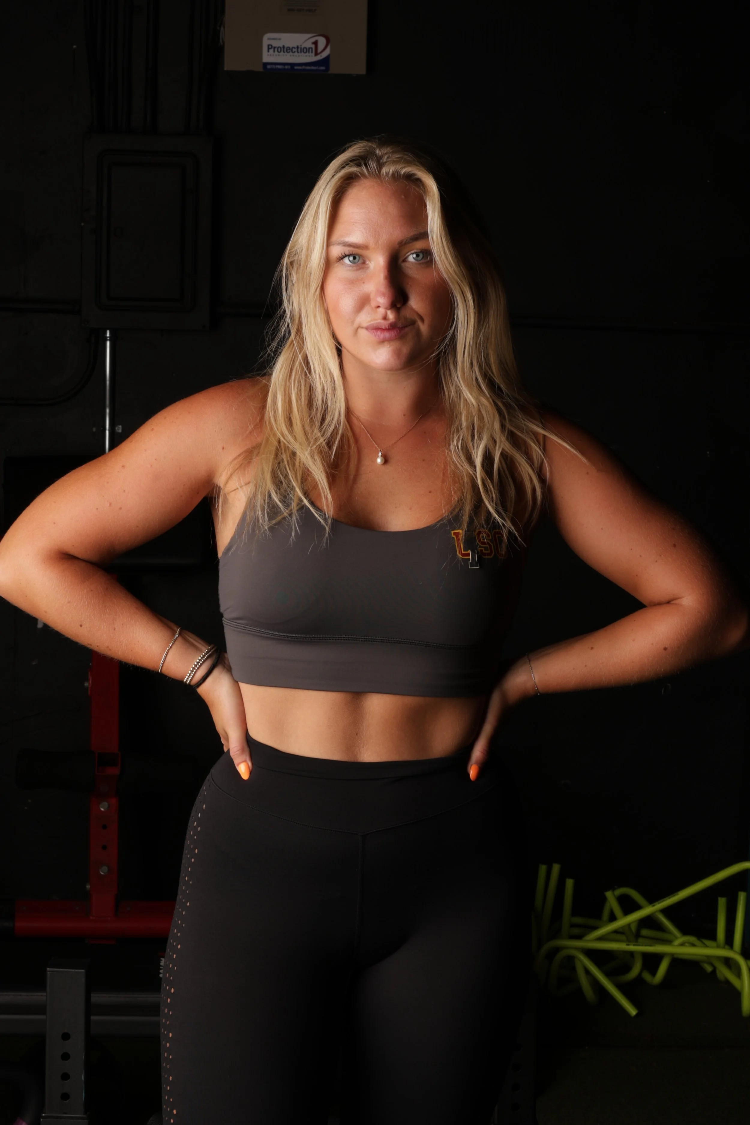 A young woman with blonde hair in workout attire posing with hands on hips in a gym.