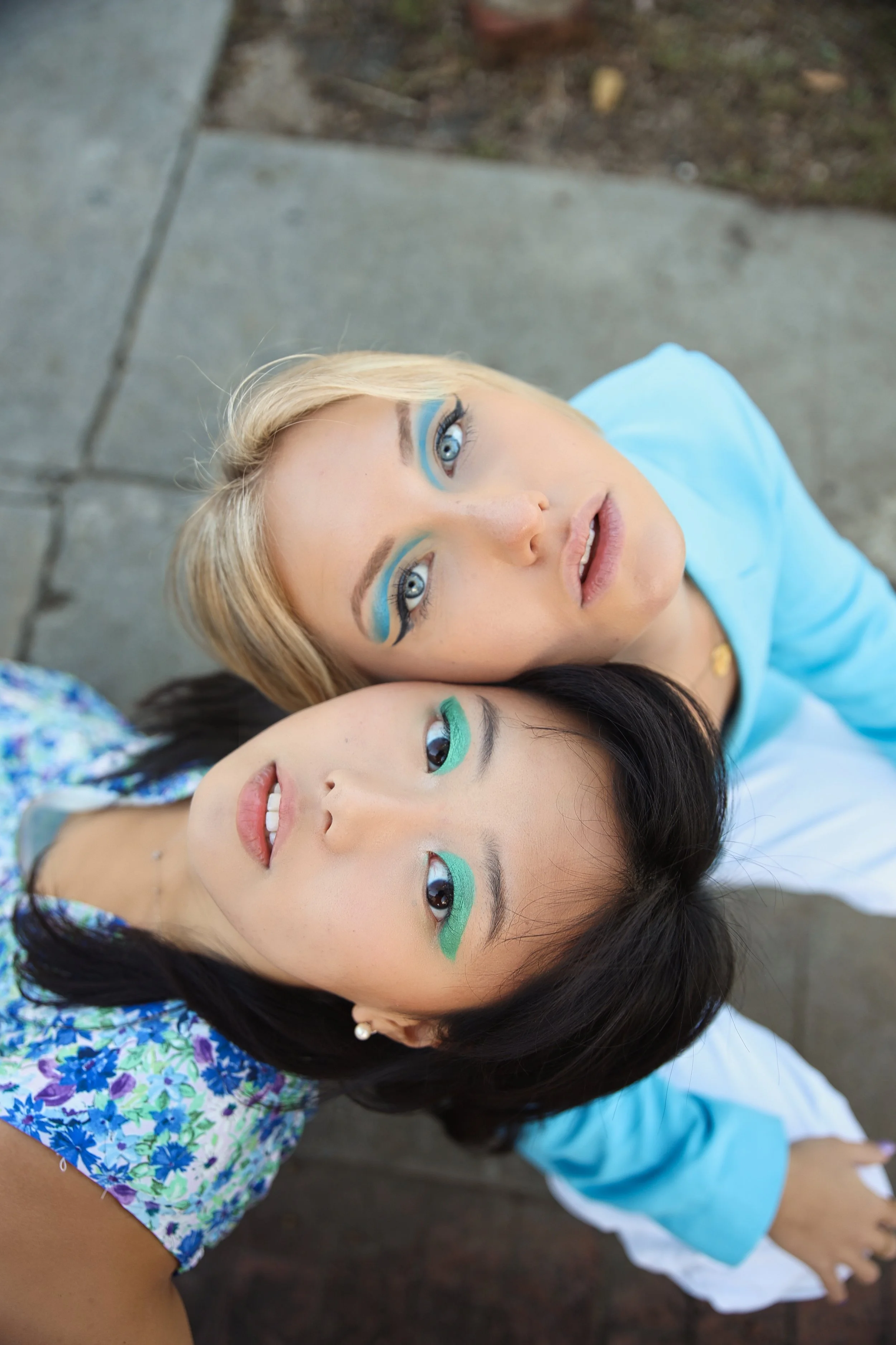 Two women lying on the ground, looking up at the camera. They have bold, colorful eye makeup with blue and green shades, and are wearing casual clothing.