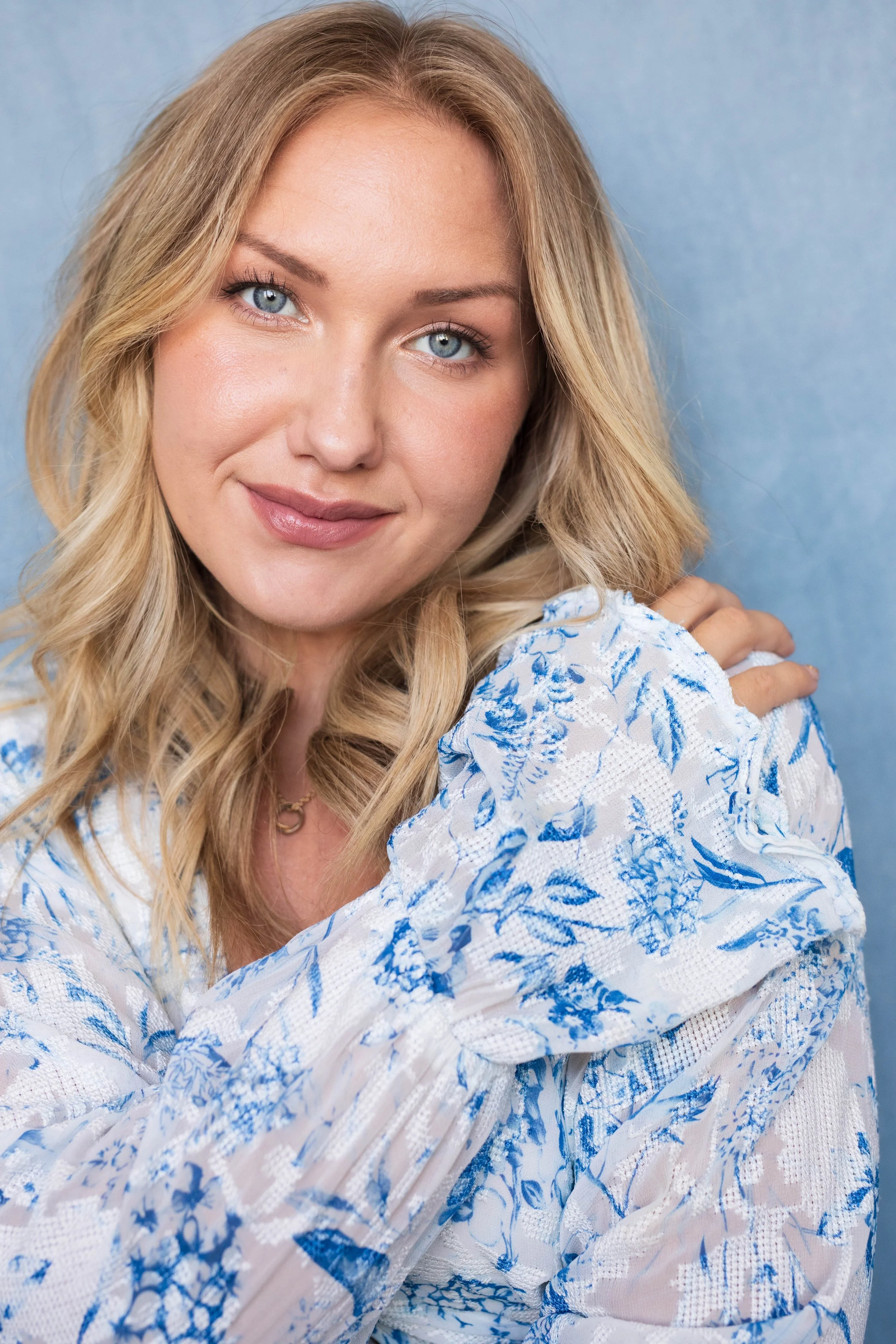 A close-up portrait of a woman with blonde hair, blue eyes, and light makeup, wearing a white blouse with blue floral patterns, against a light blue background.