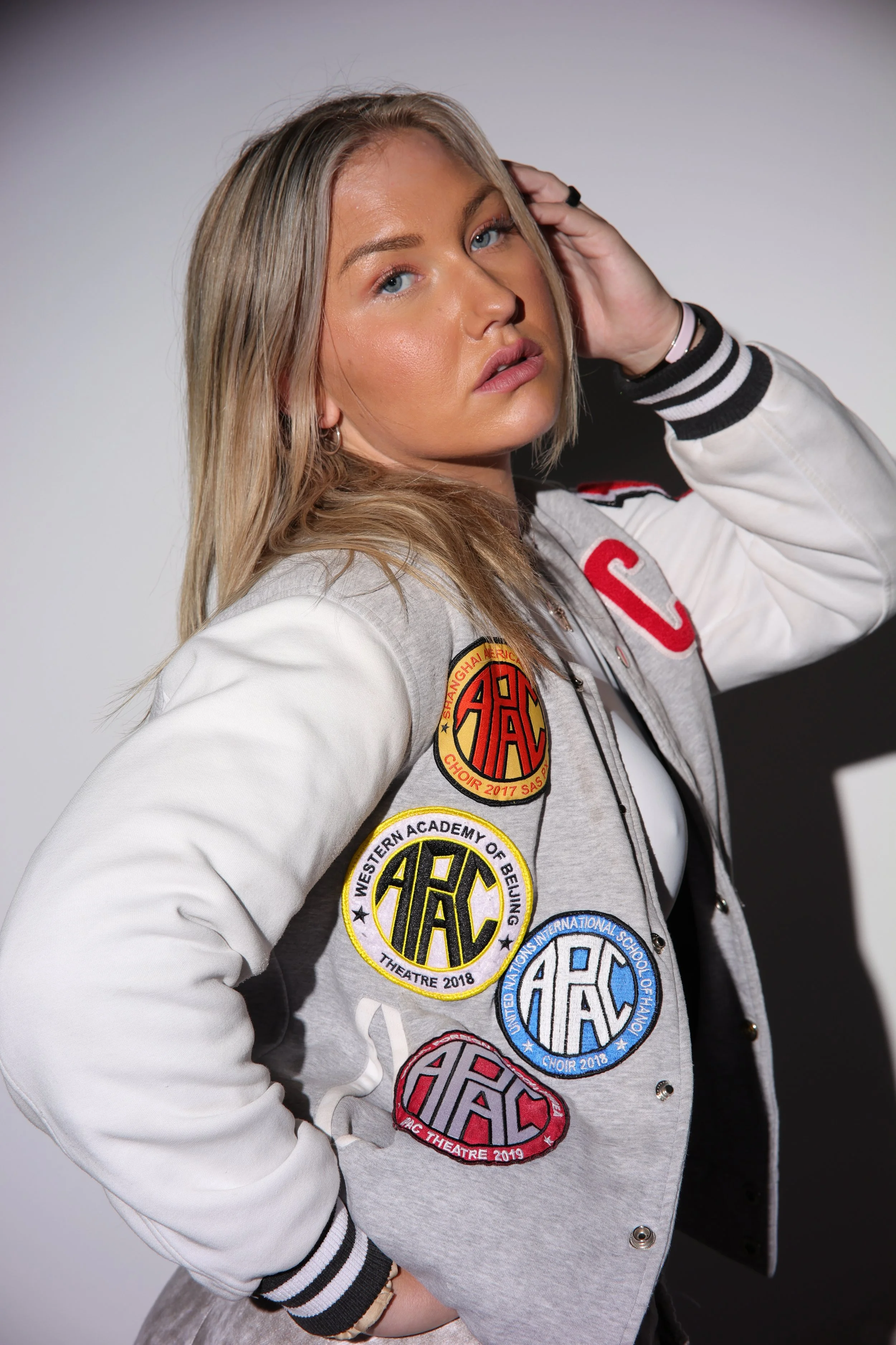 Young woman with blond hair wearing a varsity jacket decorated with patches and standing against a plain background.