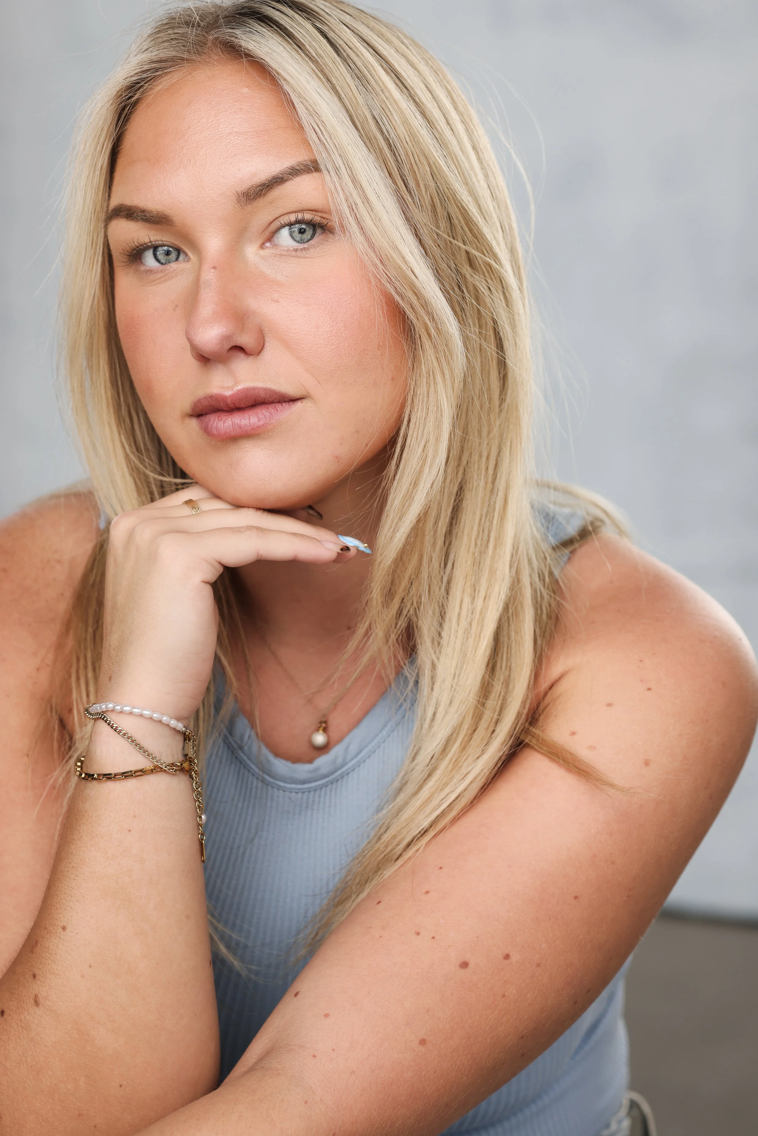 Close-up portrait of a young blonde woman with blue eyes, wearing a light blue sleeveless top, with her chin resting on her hand, looking directly at the camera.