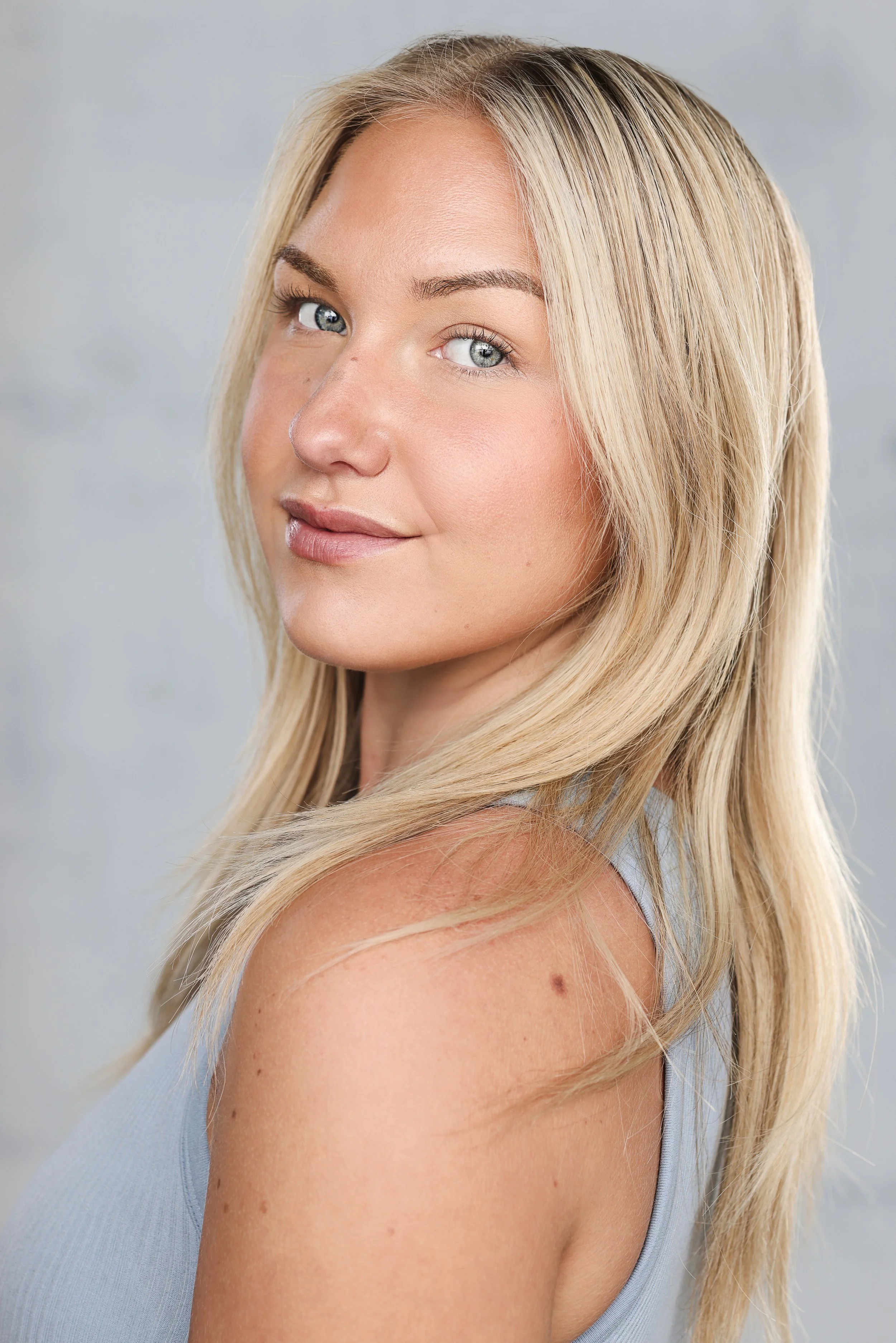 Close-up portrait of a blonde woman with blue eyes, smiling slightly, wearing a sleeveless light blue top, with a light gray background.