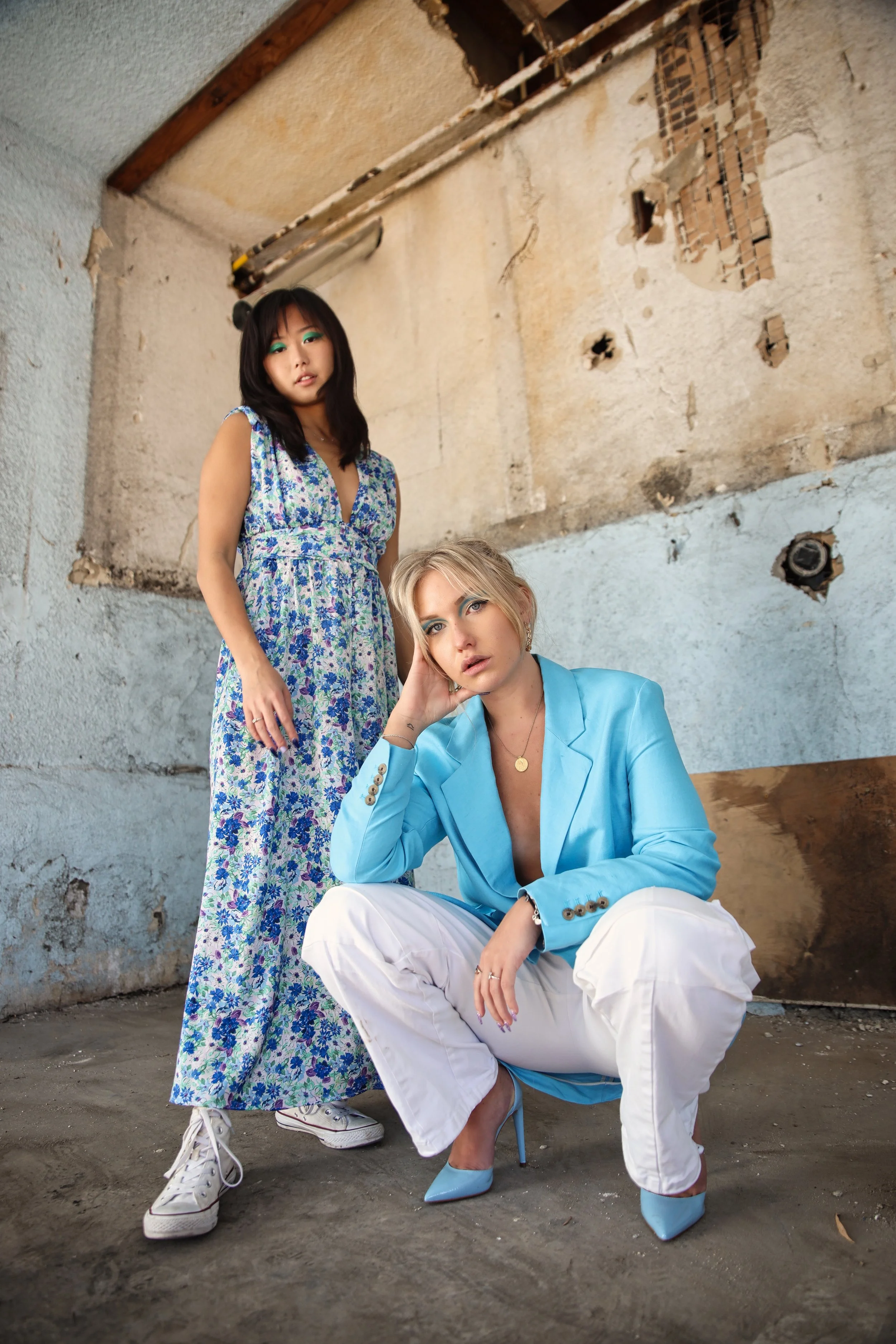 Two women in colorful fashion posing in an urban, dilapidated setting. One stands wearing a floral maxi dress with sneakers, the other crouches in a blue blazer, white pants, and high heels, with a rough, unfinished wall in the background.