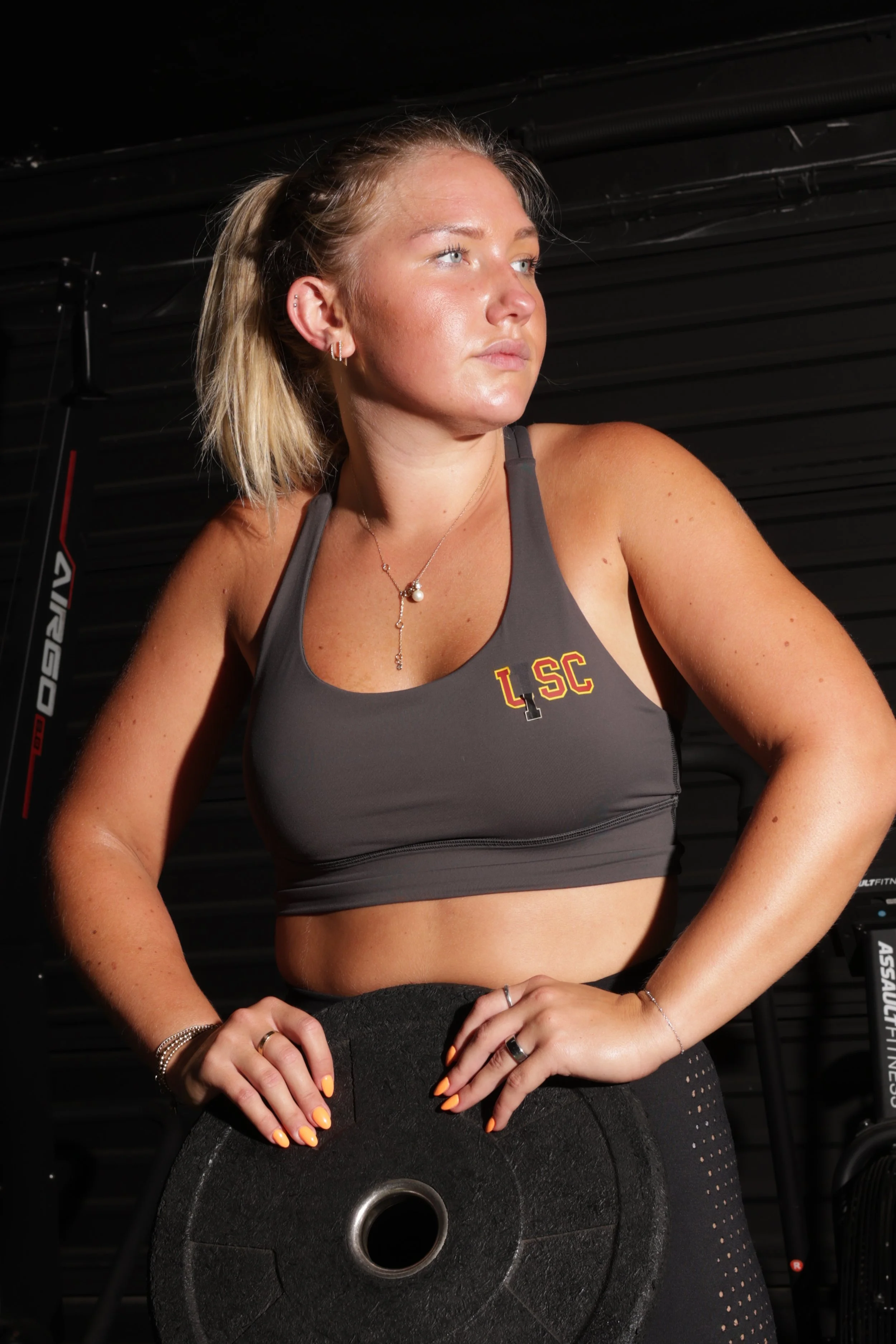 A woman with blonde hair in a high ponytail, wearing a sports bra with 'LSC' embroidery, holding a weight plate in a gym setting.