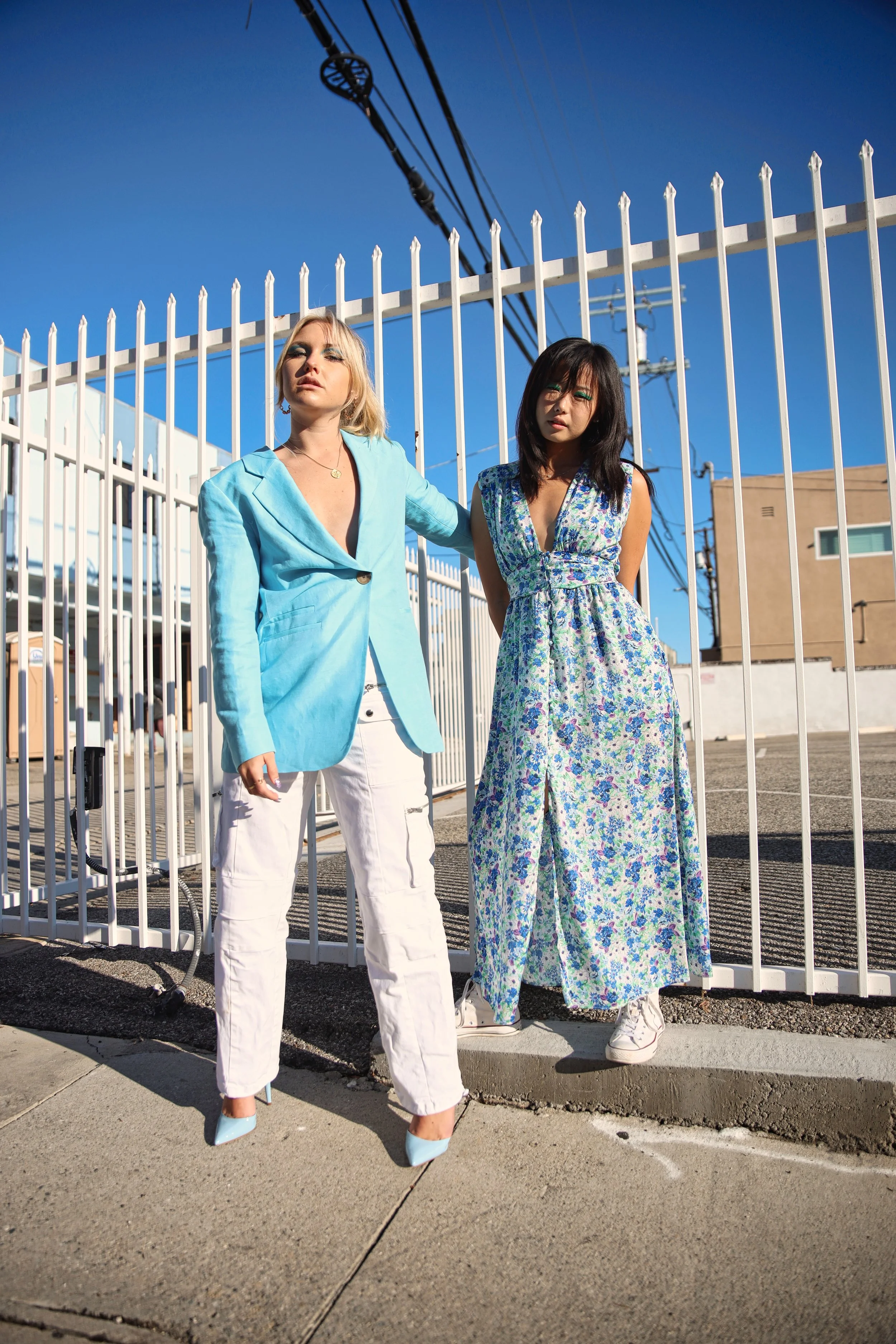 Two women standing outdoors in front of a white fence, wearing stylish clothes, with clear blue sky above and power lines overhead.