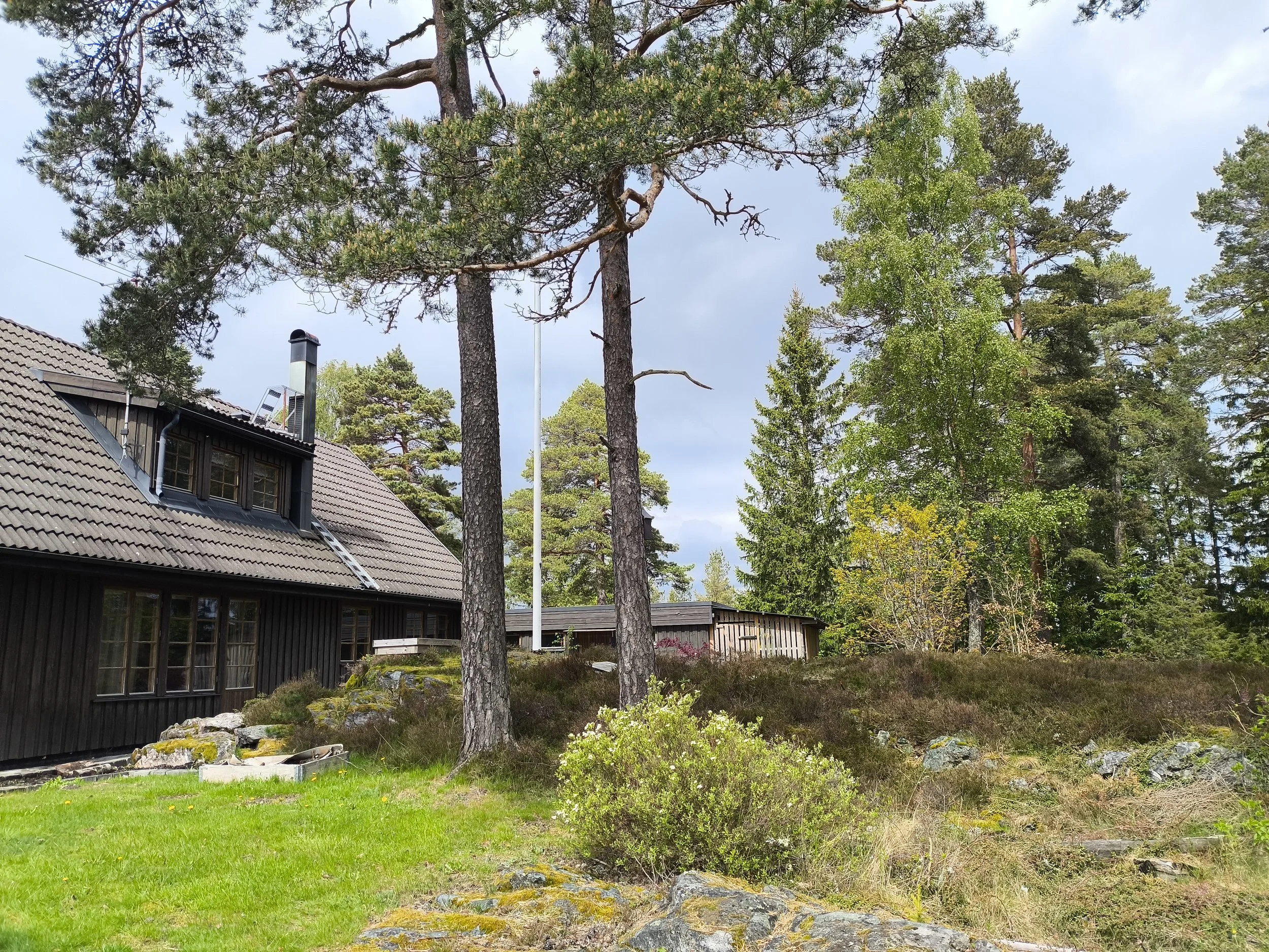A house with a sloped roof and a chimney, surrounded by tall pine trees and greenery, under a partly cloudy sky. West Cottage Sweden, Åsensbruk.